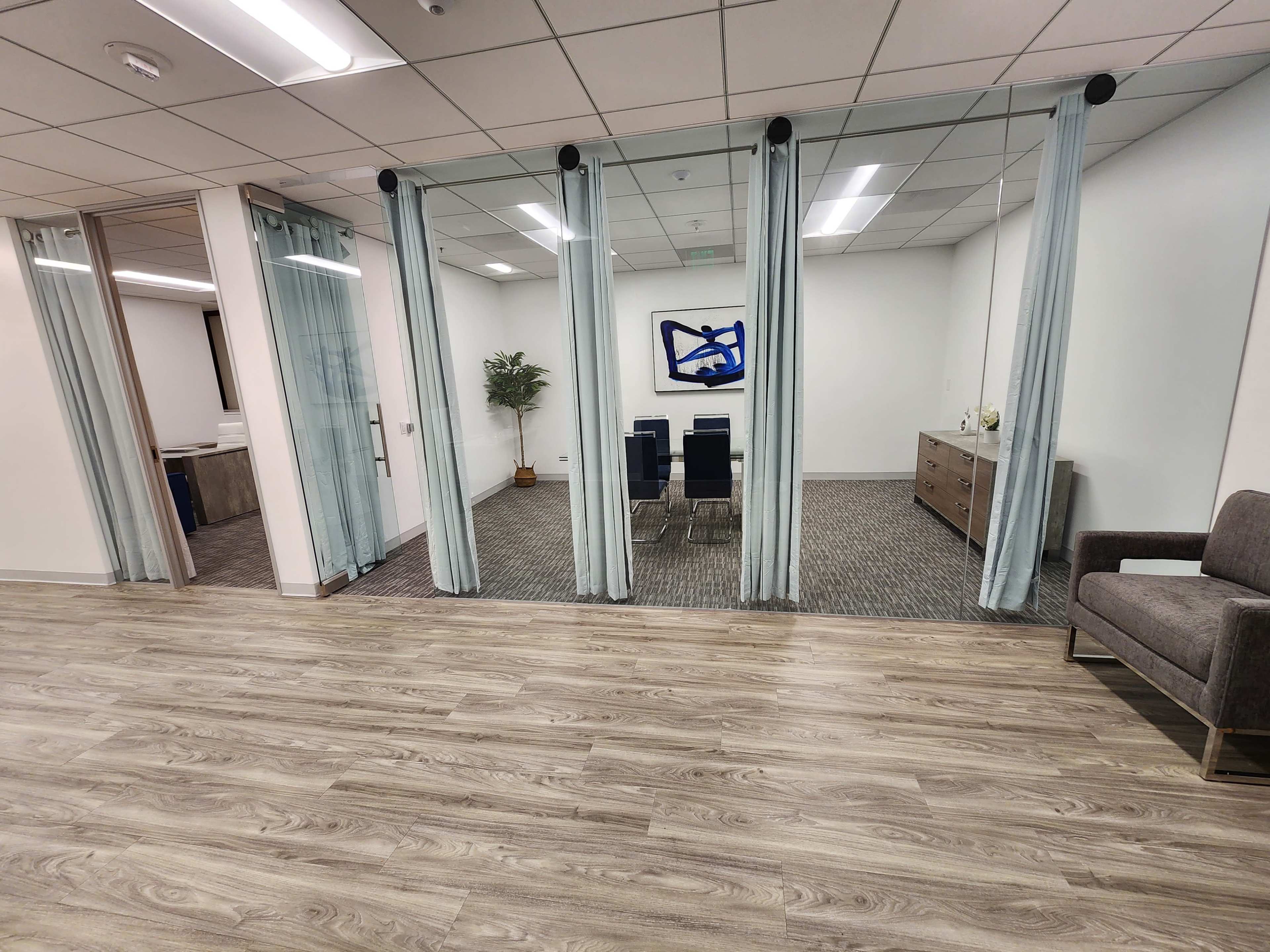 The image shows a modern office space with transparent partitions, a seating area, and a small conference room visible through the glass.