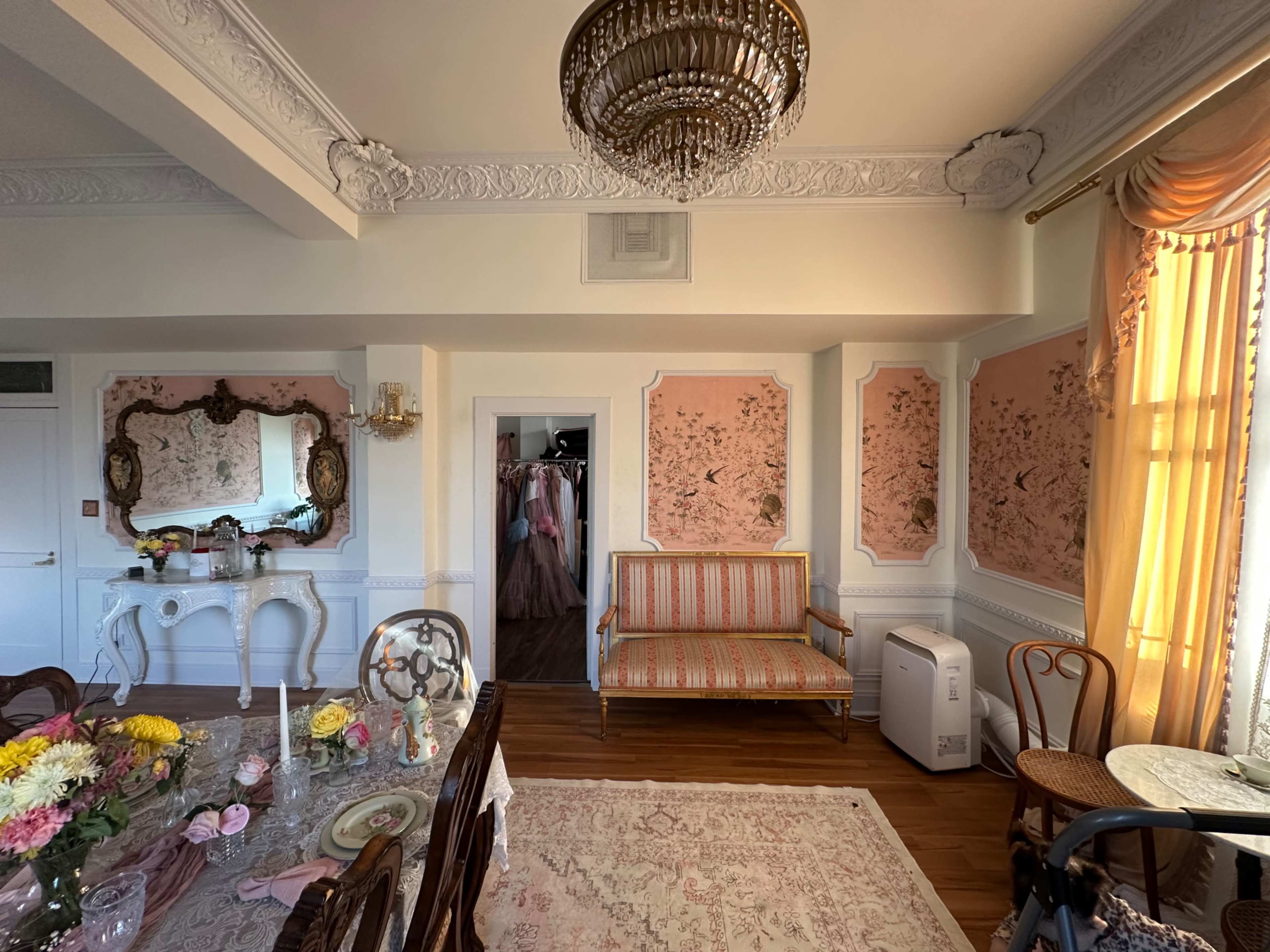 The image shows a well-decorated room featuring floral wallpaper, a chandelier, a dining table with flowers, and a vintage sofa, with a closet area visible in the background.