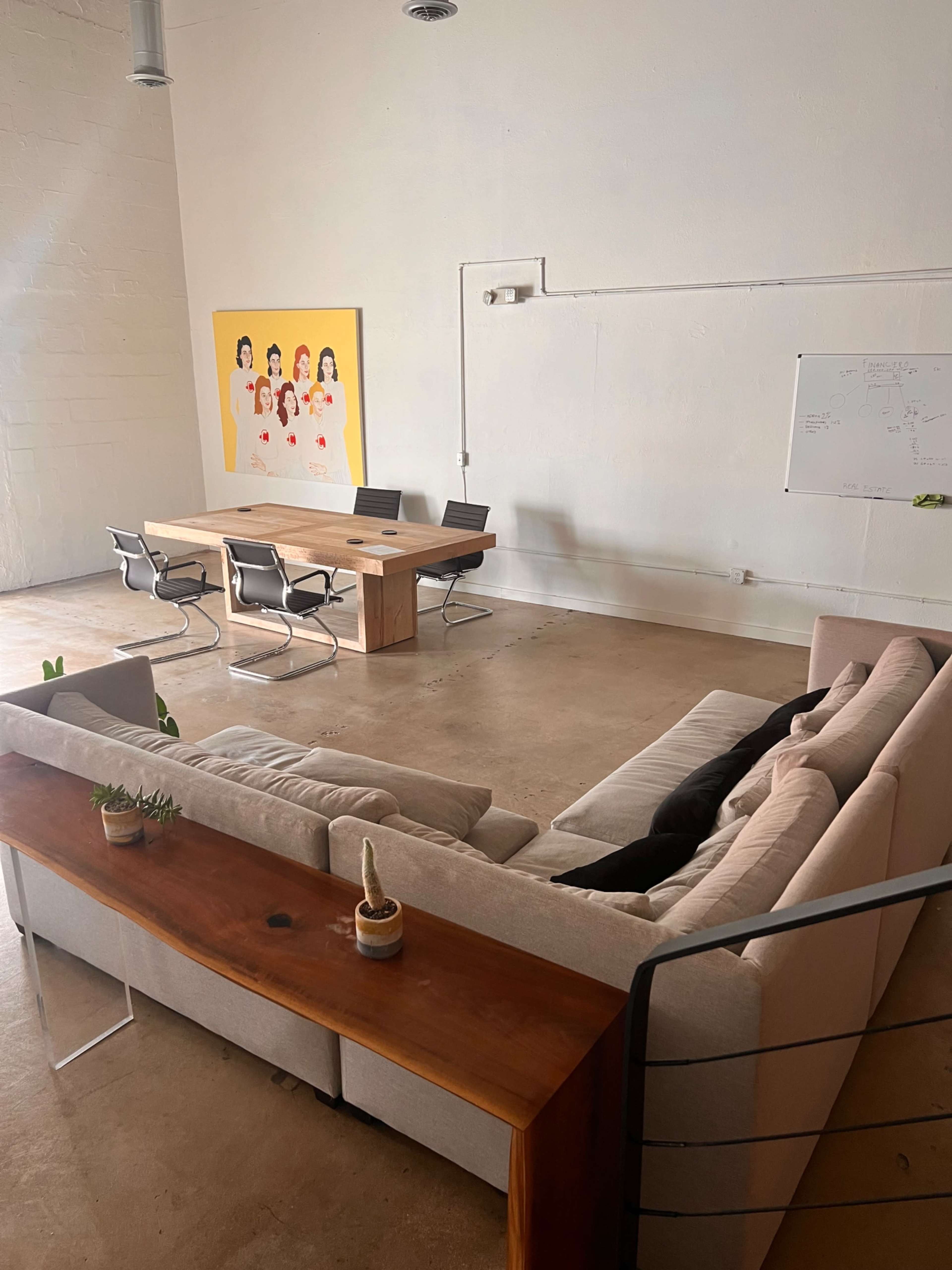 A modern meeting room features a large wooden table surrounded by chairs, with a sectional sofa and a colorful artwork on the wall.