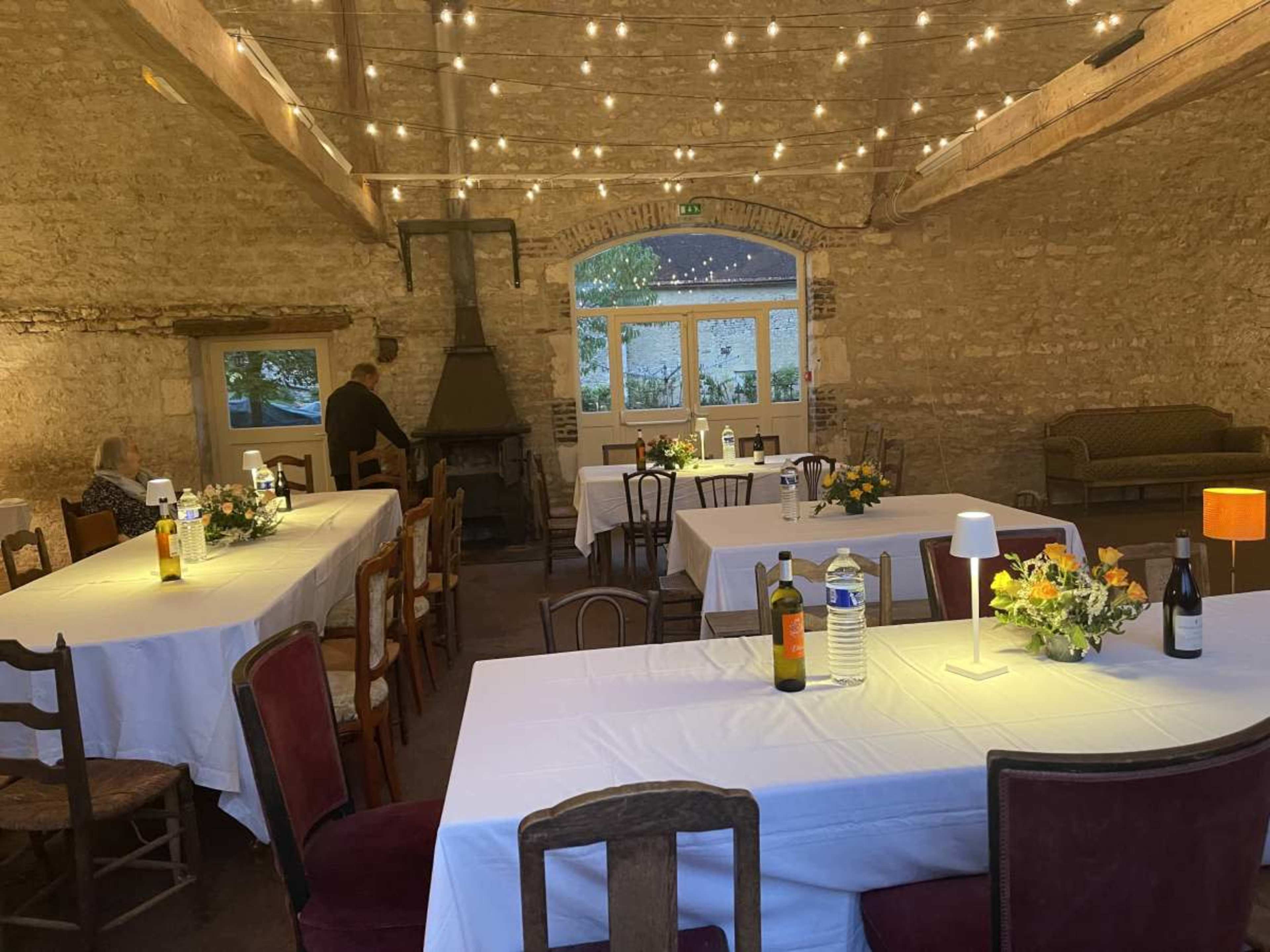 The image shows a rustic interior space featuring several tables with white tablecloths, a fireplace, and string lights overhead, creating a cozy atmosphere.
