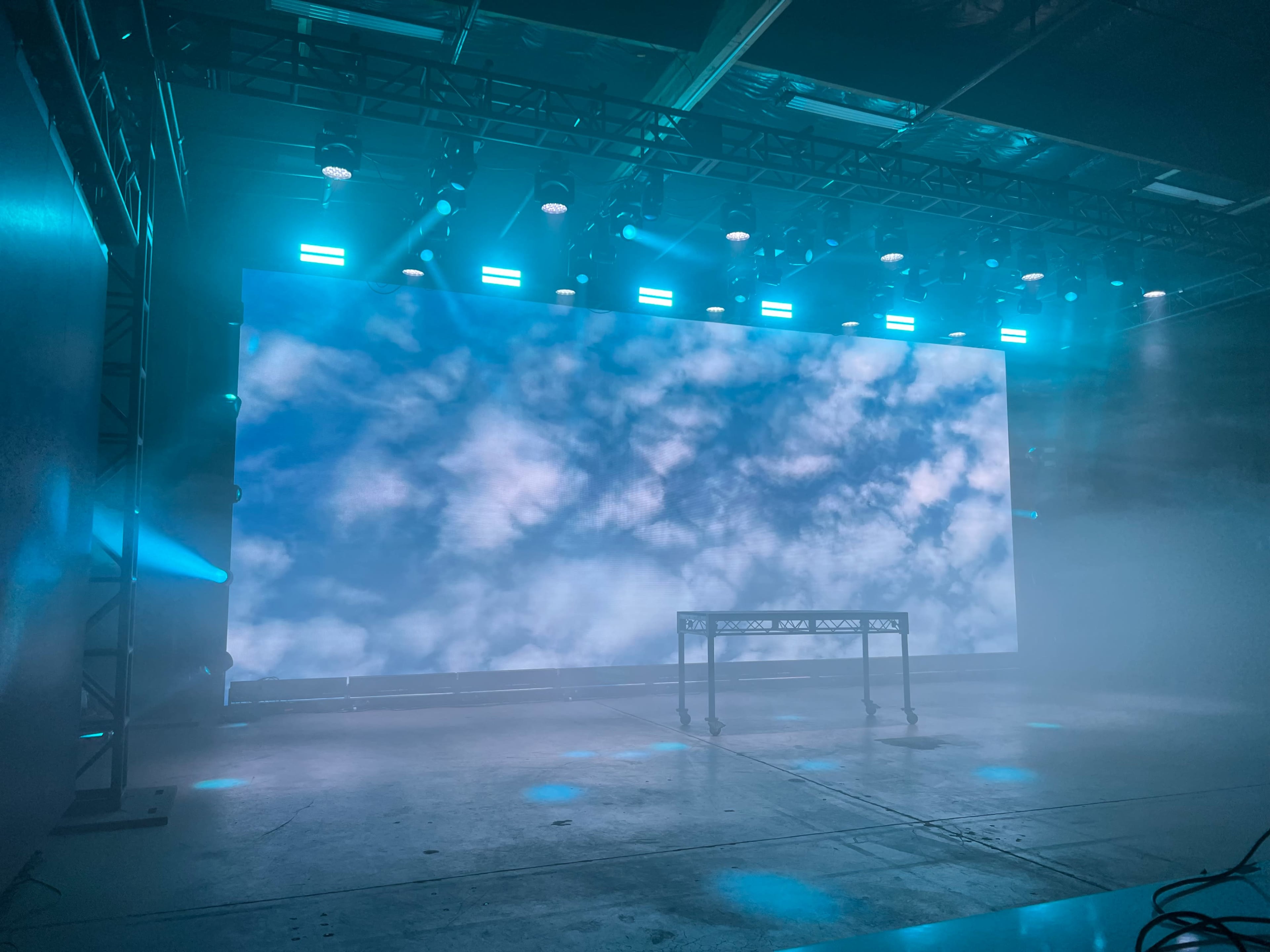 A large screen displaying cloud imagery is illuminated by blue stage lights in a performance venue.