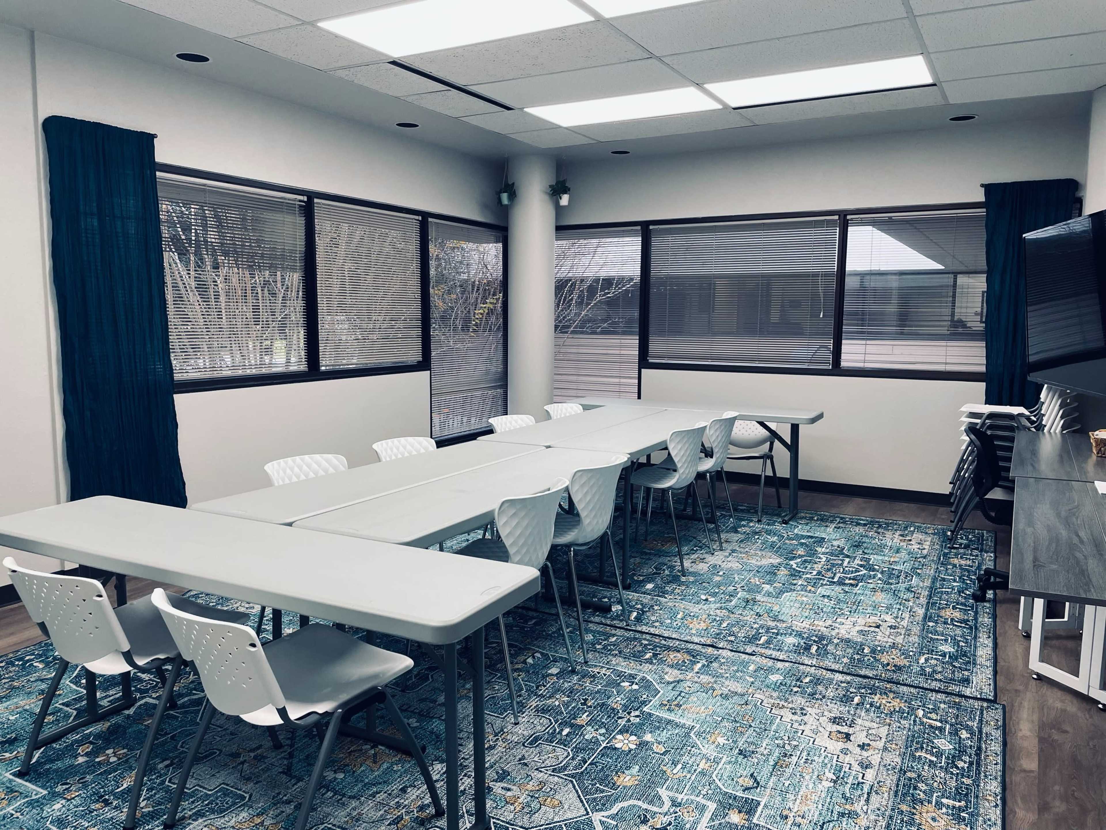 A conference room features multiple tables arranged in a U-shape, surrounded by chairs, with large windows allowing natural light to enter.