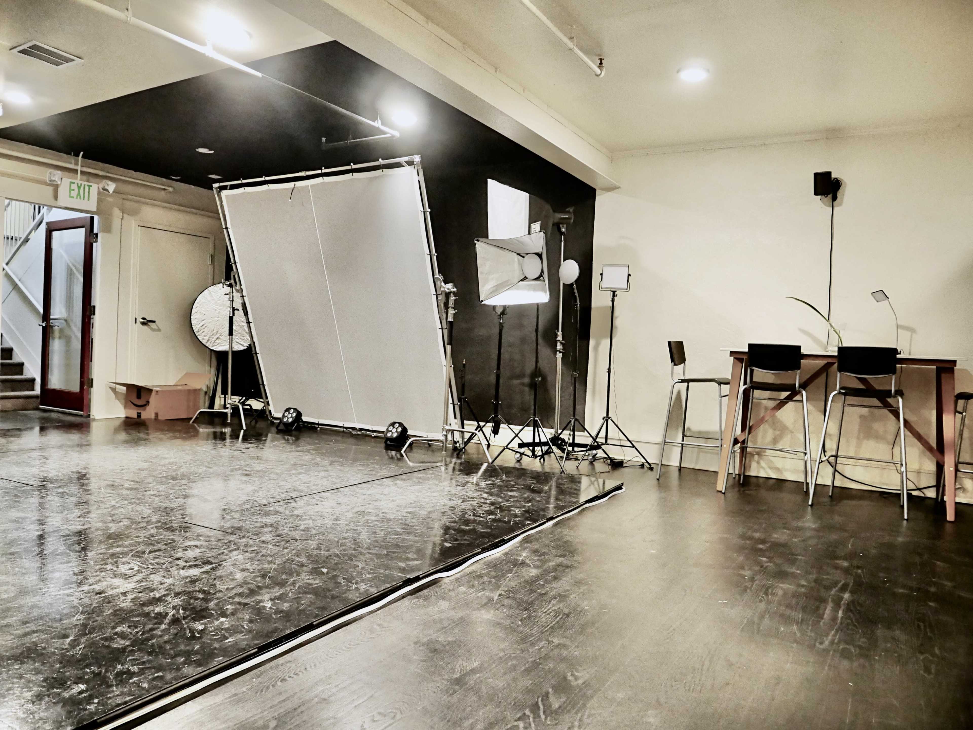 The image shows an empty studio space with a large backdrop, lighting equipment, and a table with chairs set against a black wall.