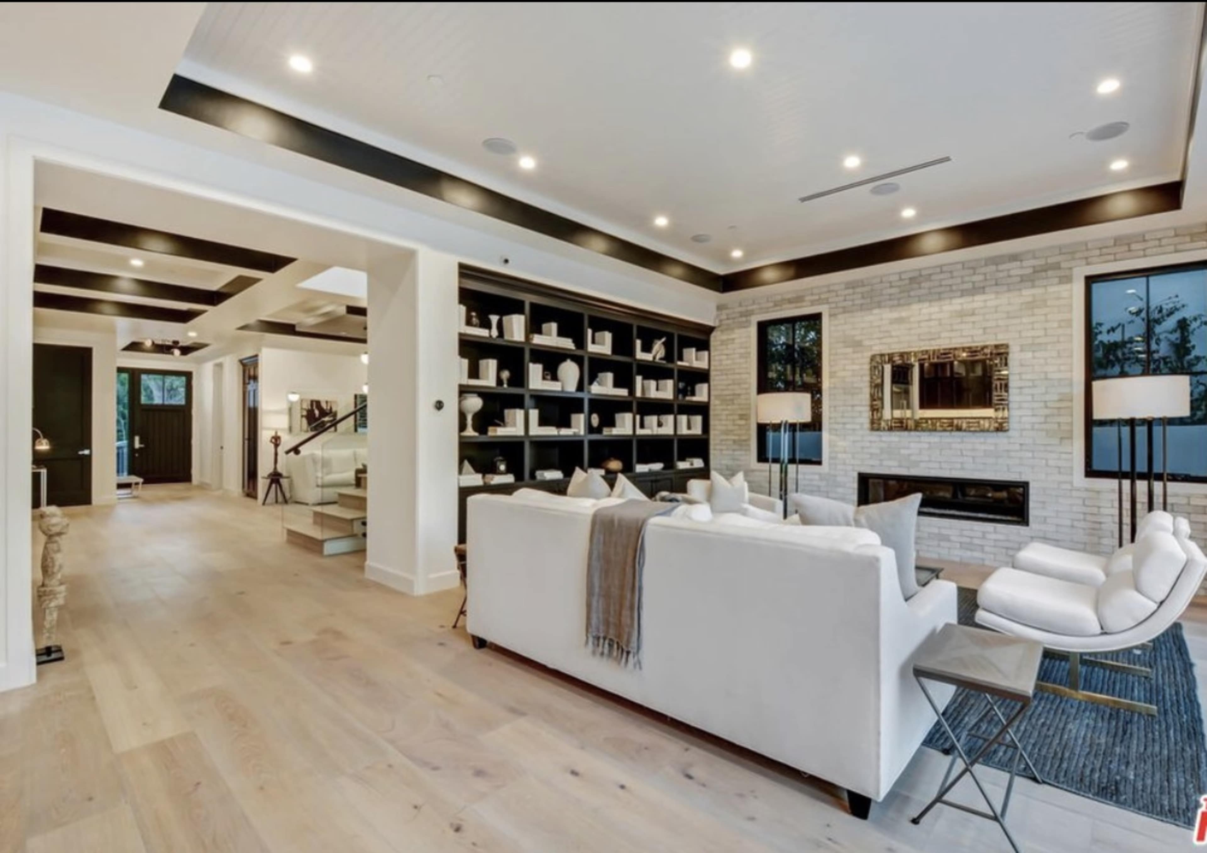 The image shows a modern living room with white furniture, a fireplace, and large windows, featuring a minimalist bookshelf and light wood flooring.