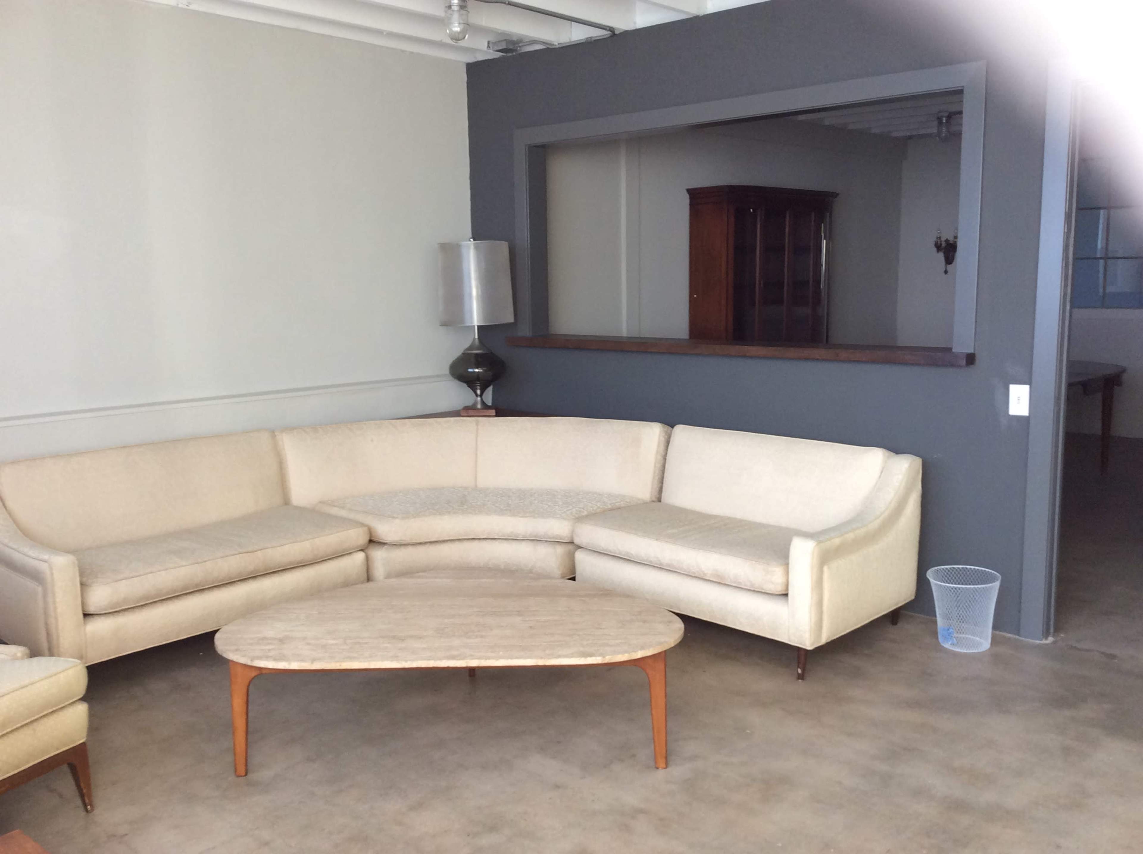 The image shows a beige sectional sofa with a mid-century modern coffee table in a sparsely furnished room featuring a gray wall and a doorway leading to another space.