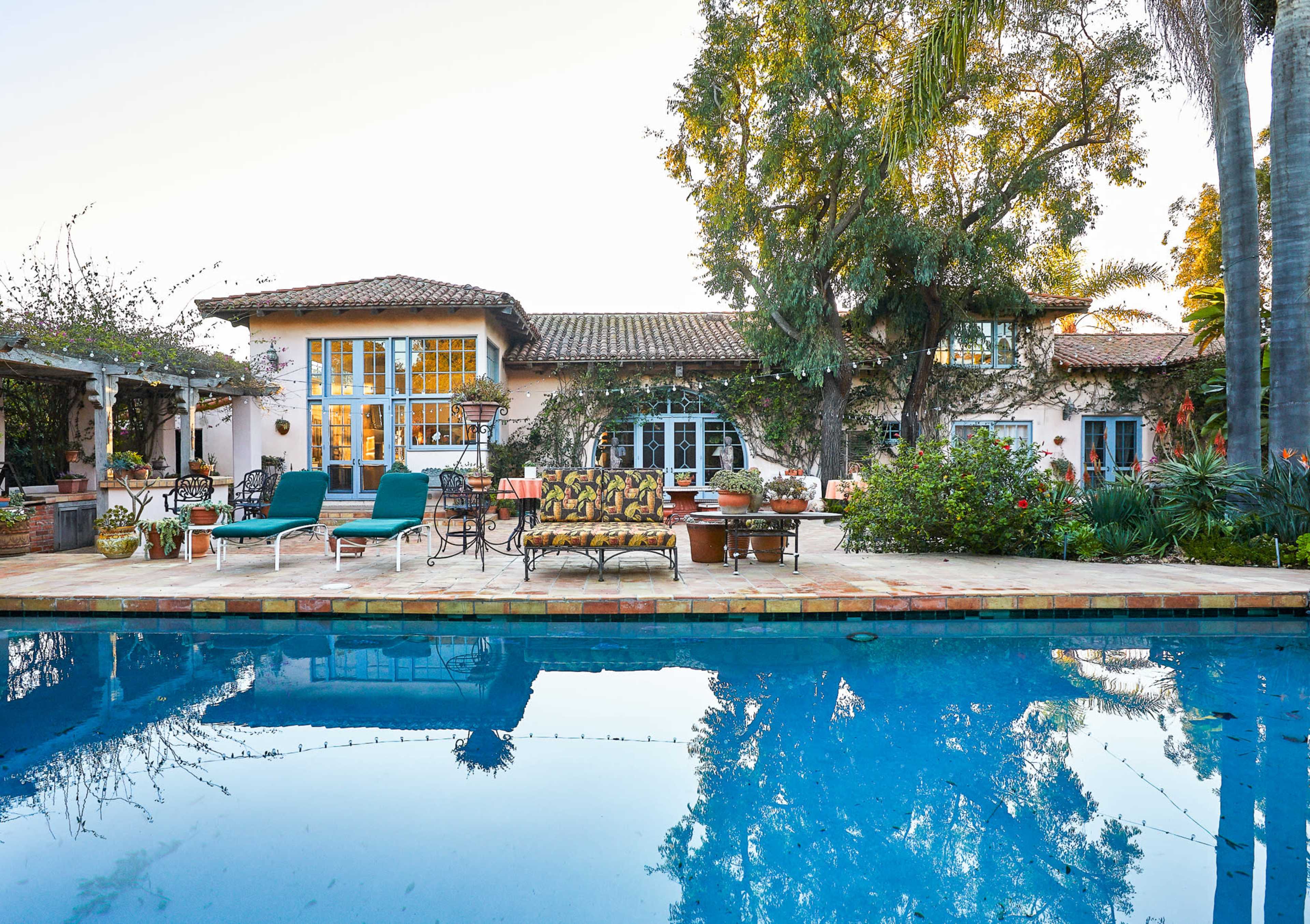 A spacious outdoor area features a swimming pool surrounded by lounge furniture and a view of a Mediterranean-style house with patio seating and lush greenery.