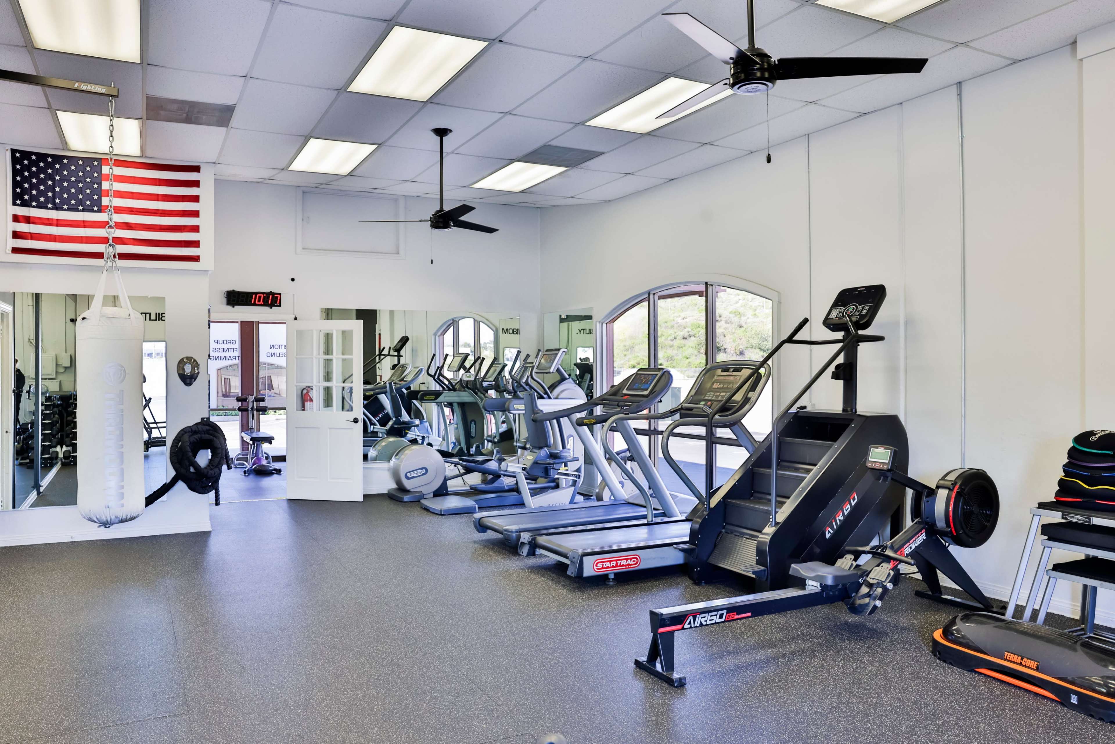The image shows an indoor gym with various cardio machines, a punching bag, and an American flag displayed on the wall.
