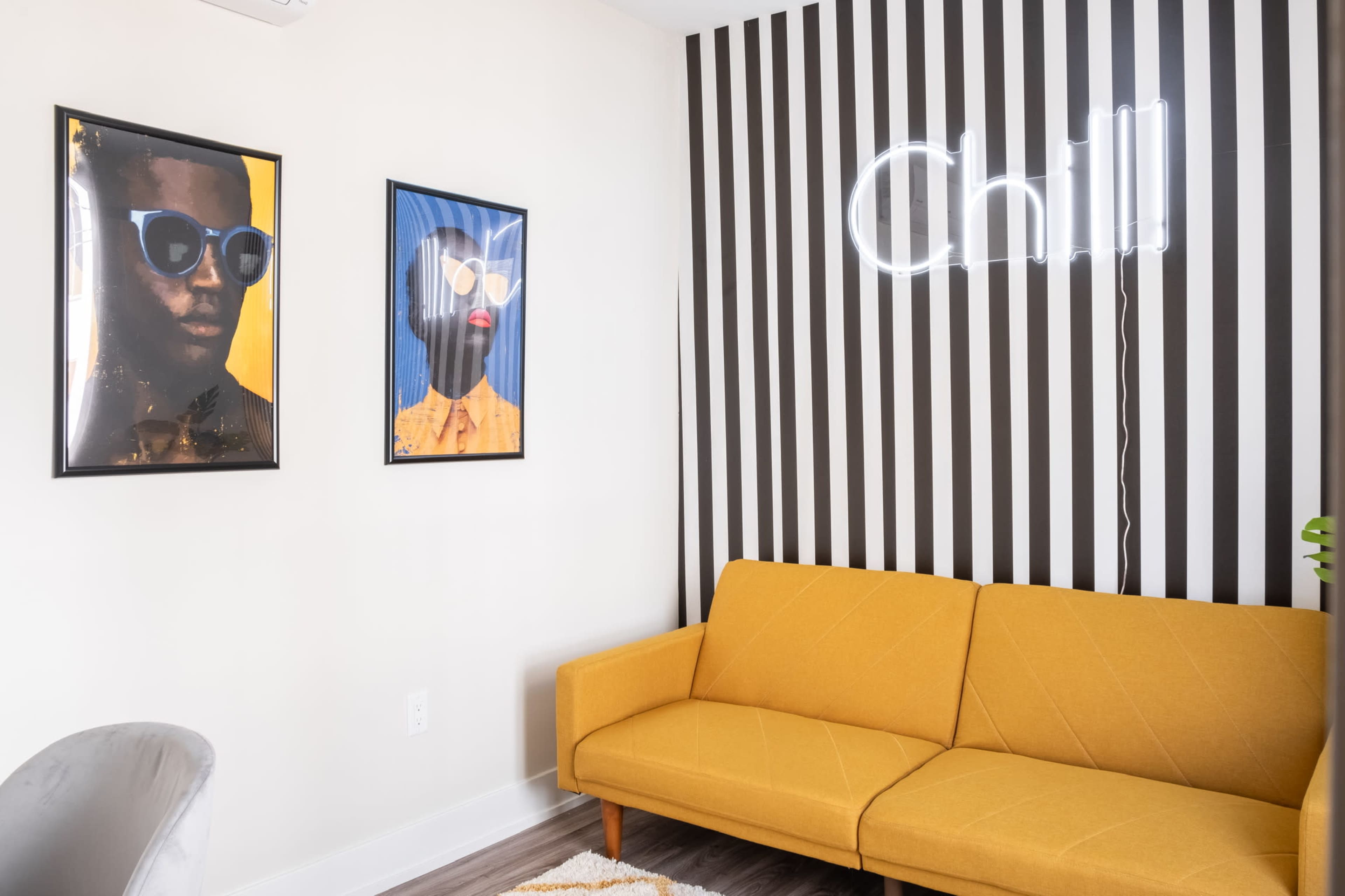 A modern lounge area with a yellow couch, two framed portraits on the wall, and a neon sign that reads "Chill" against a striped backdrop.