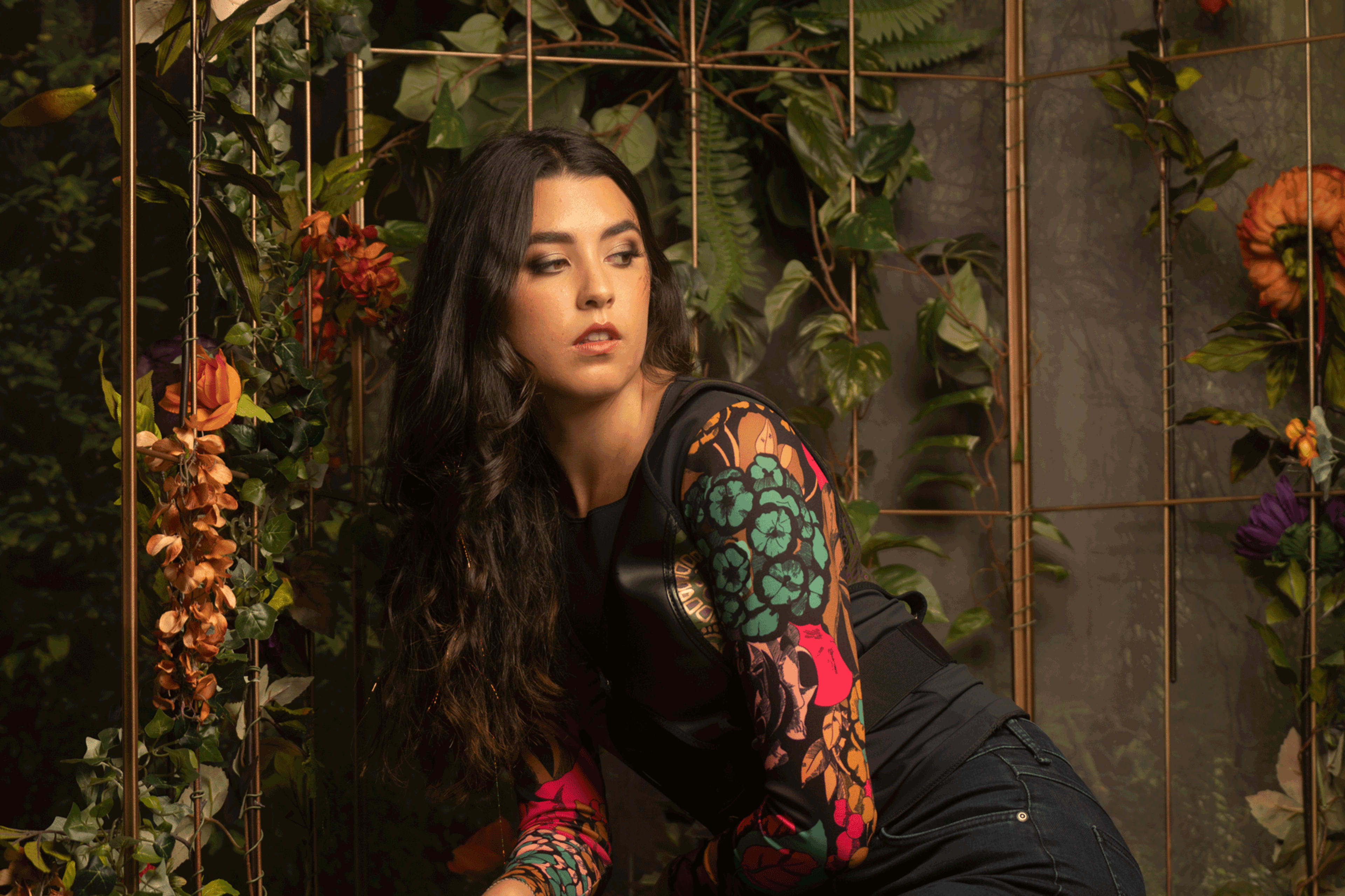 A woman with long, dark hair poses in front of a backdrop of vibrant flowers and greenery.