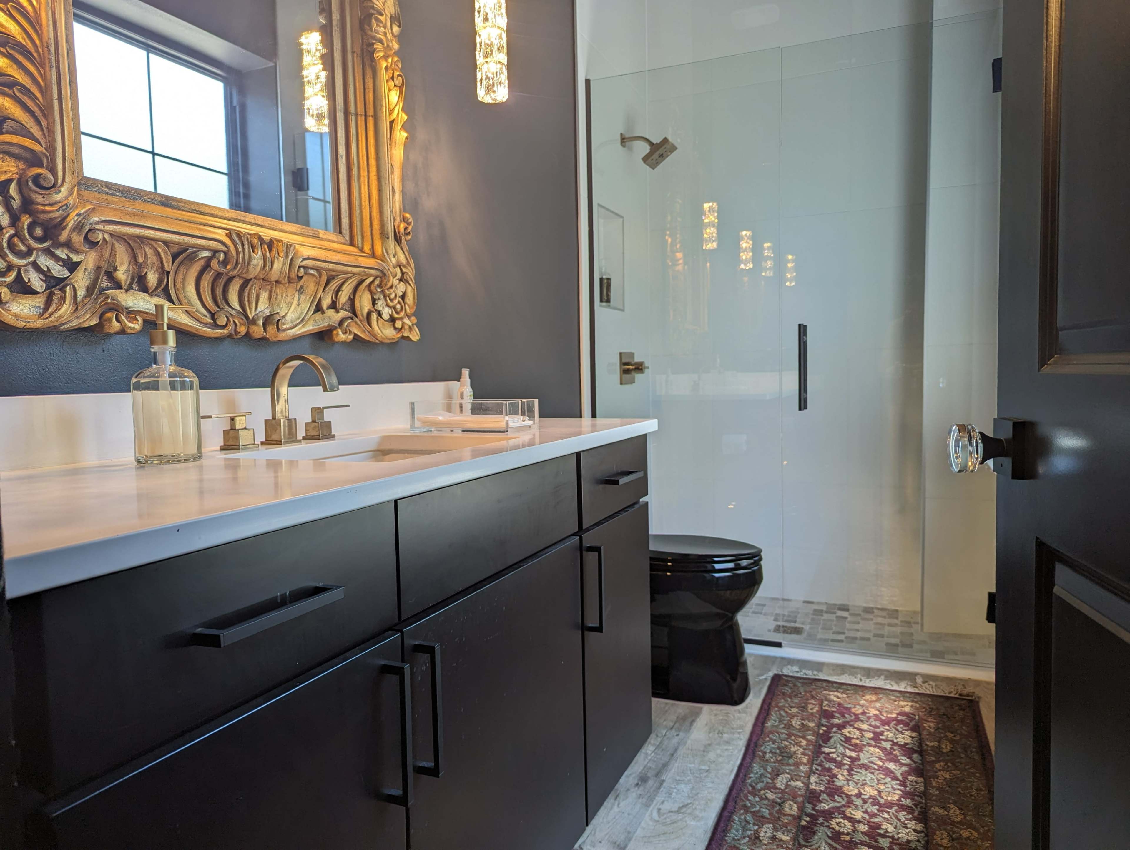 A modern bathroom featuring a black vanity with a marble countertop, a large ornate mirror, a glass shower enclosure, and a toilet.