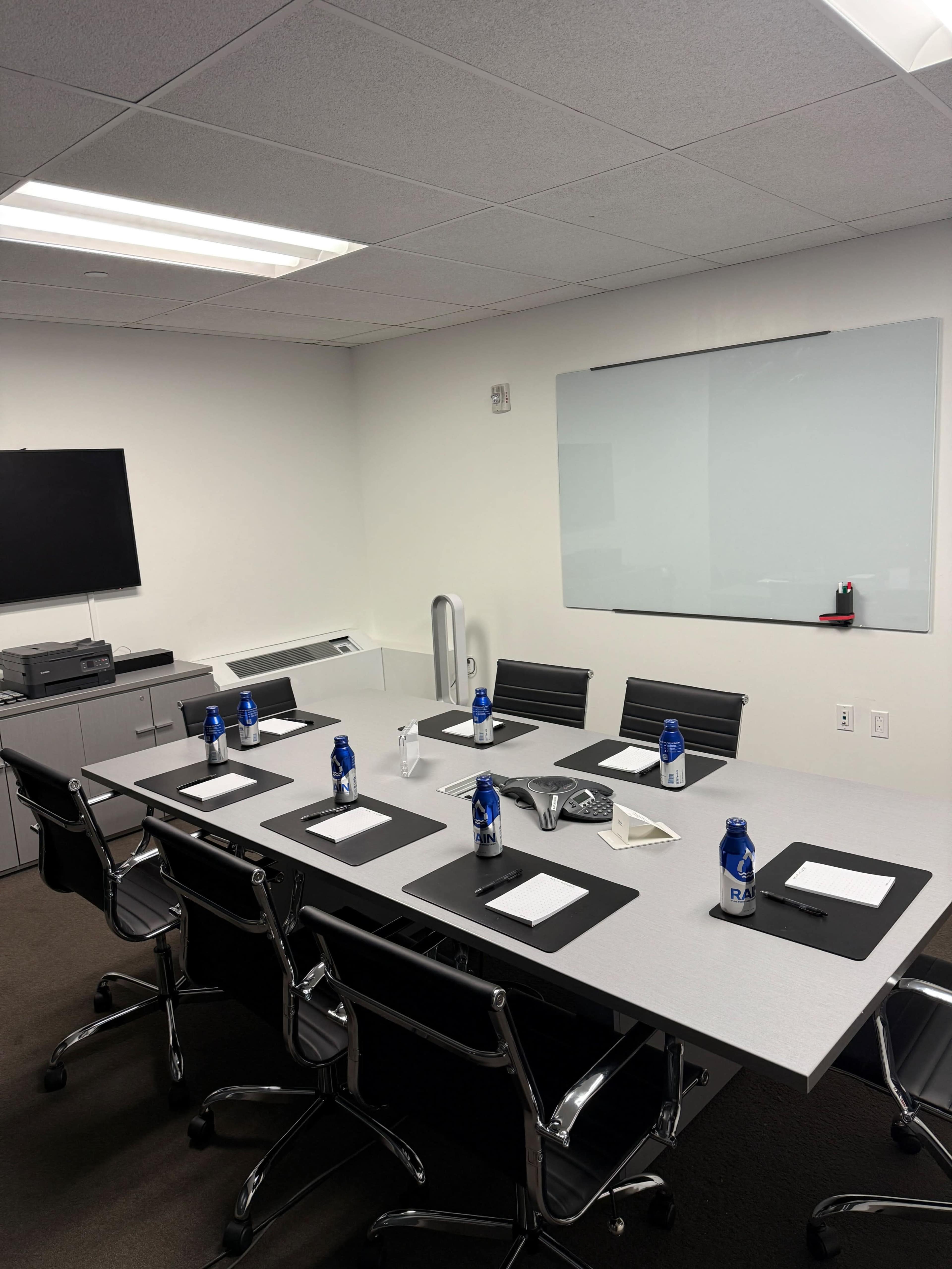 A modern conference room features a long table with black placemats, white notepads, water bottles, and a television mounted on the wall.