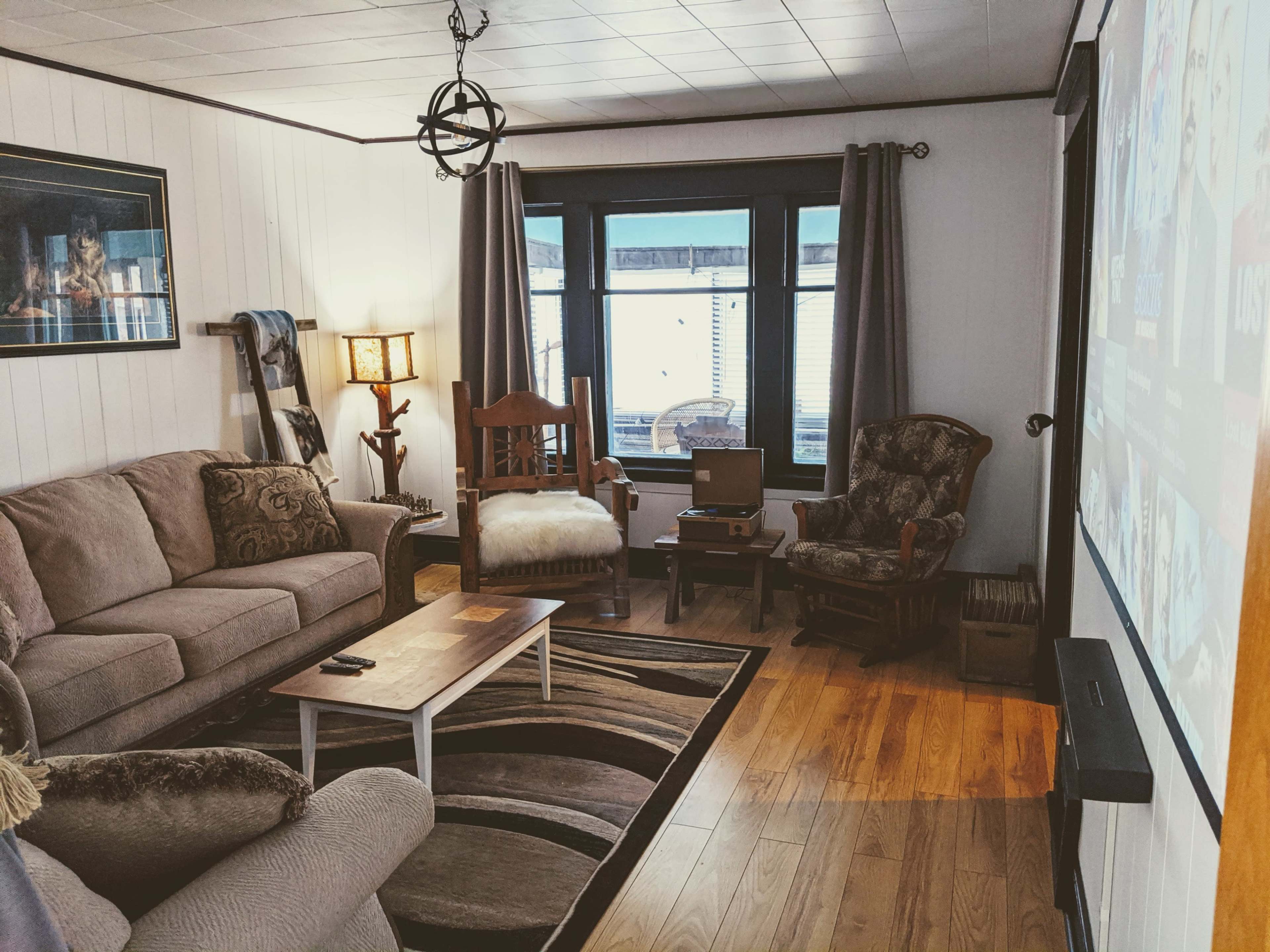 The living room features a sofa, armchairs, a coffee table, and a large projector screen against one wall, with windows letting in natural light.