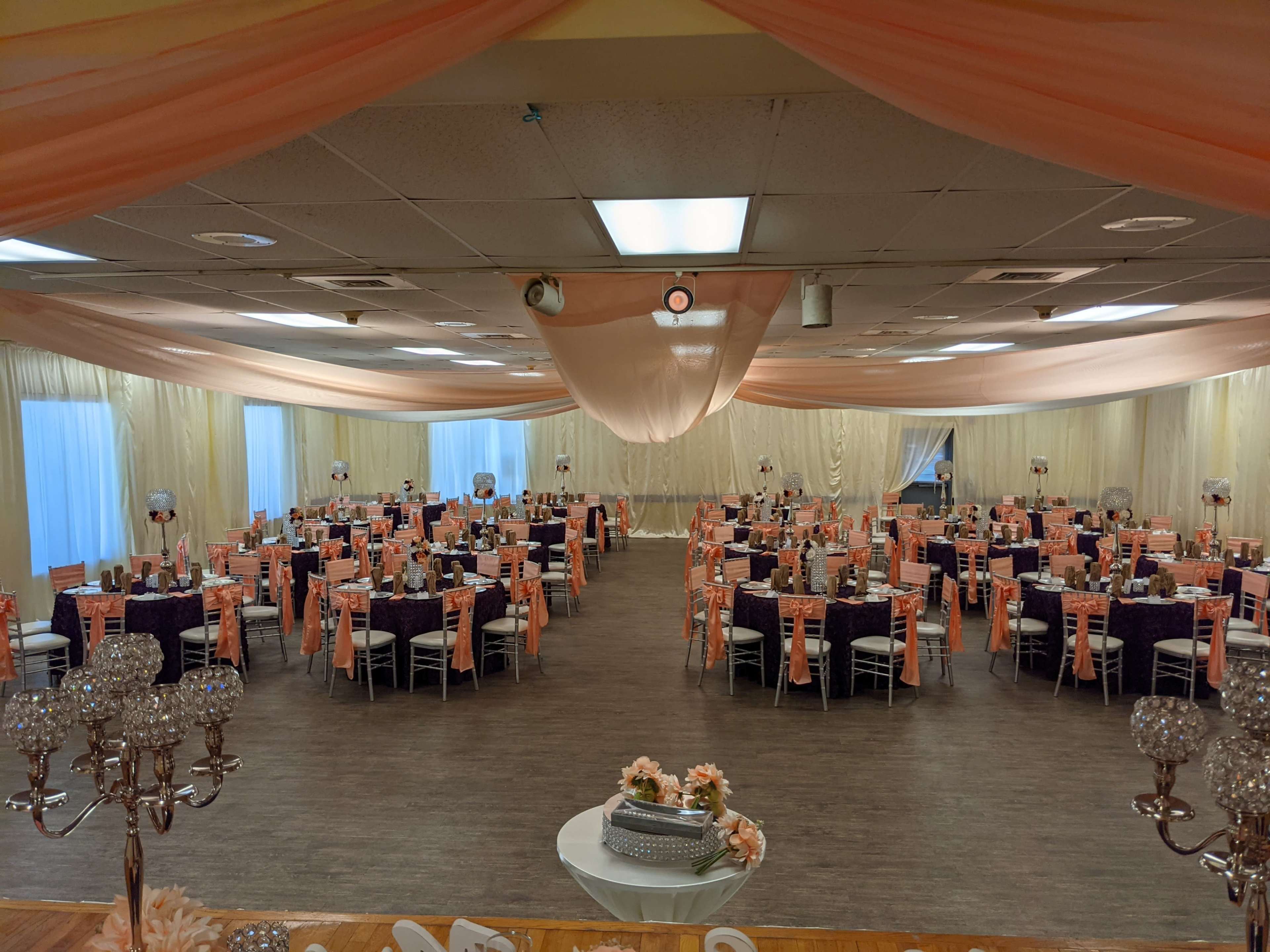 The image shows a large banquet hall with round tables set for an event, each covered with dark purple cloths and adorned with pink and white decorations.