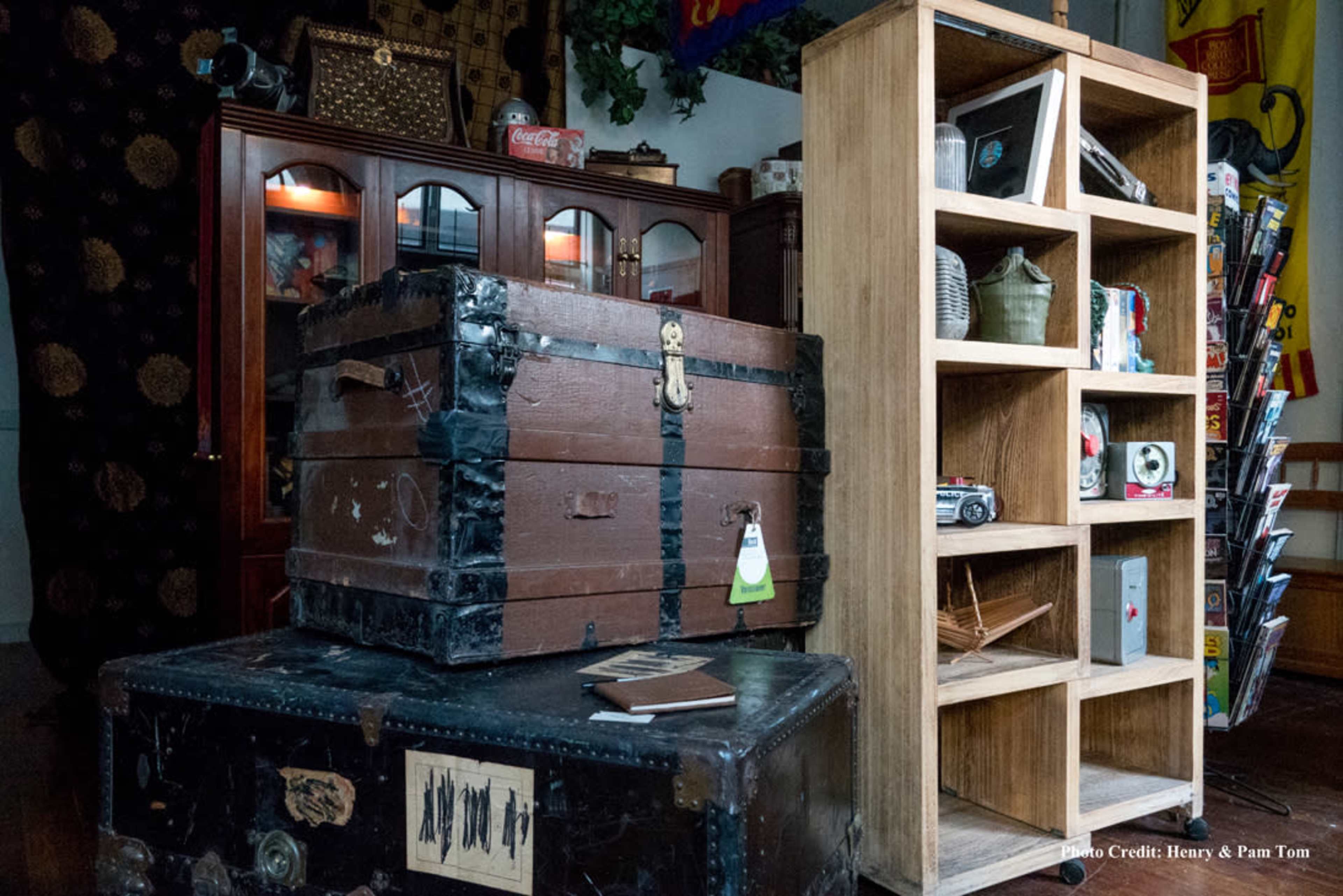 A wooden chest with black straps resting on a table beside a tall wooden shelf filled with various items.