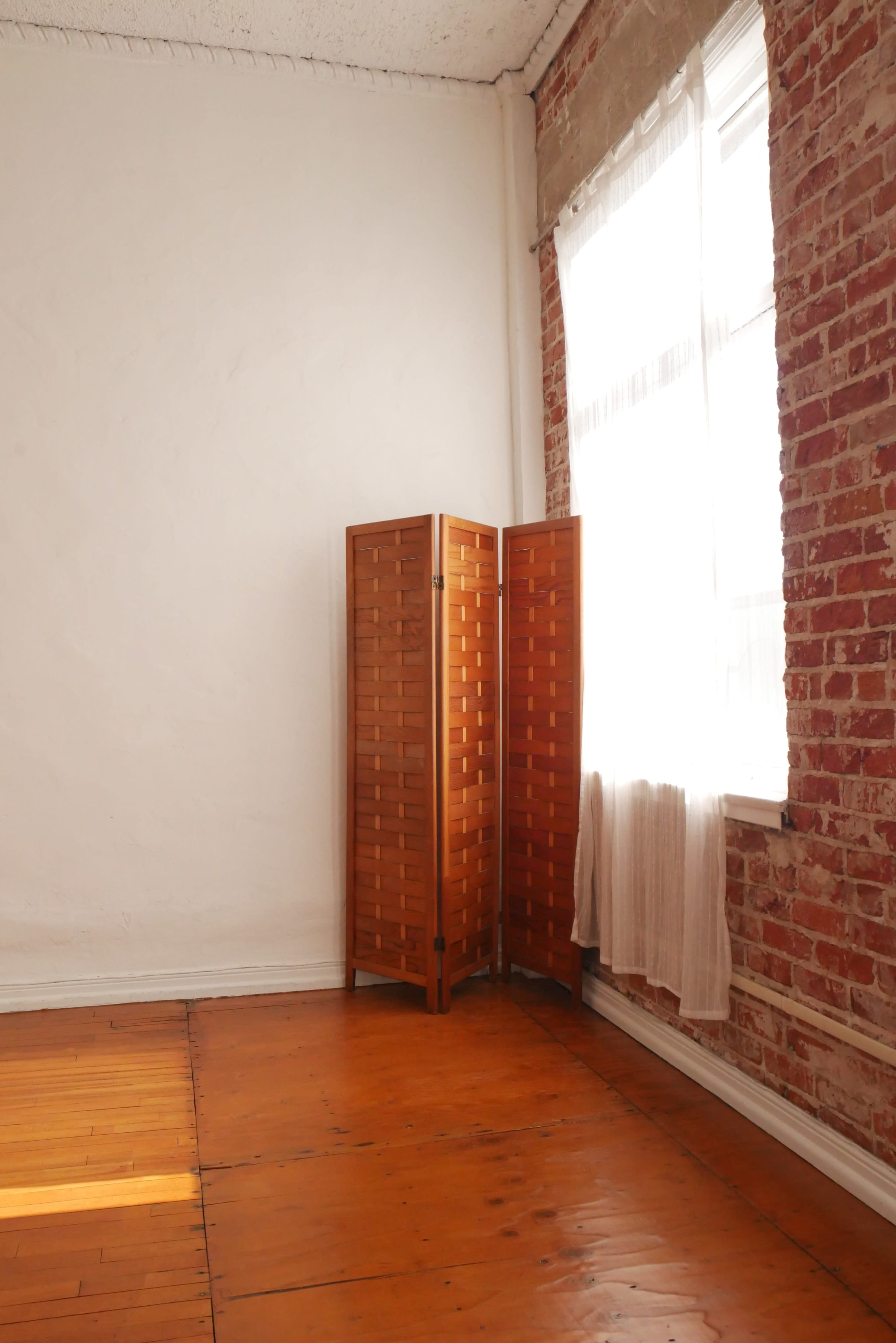 A wooden folding screen stands in the corner of a room with exposed brick walls and a window covered by a light curtain.