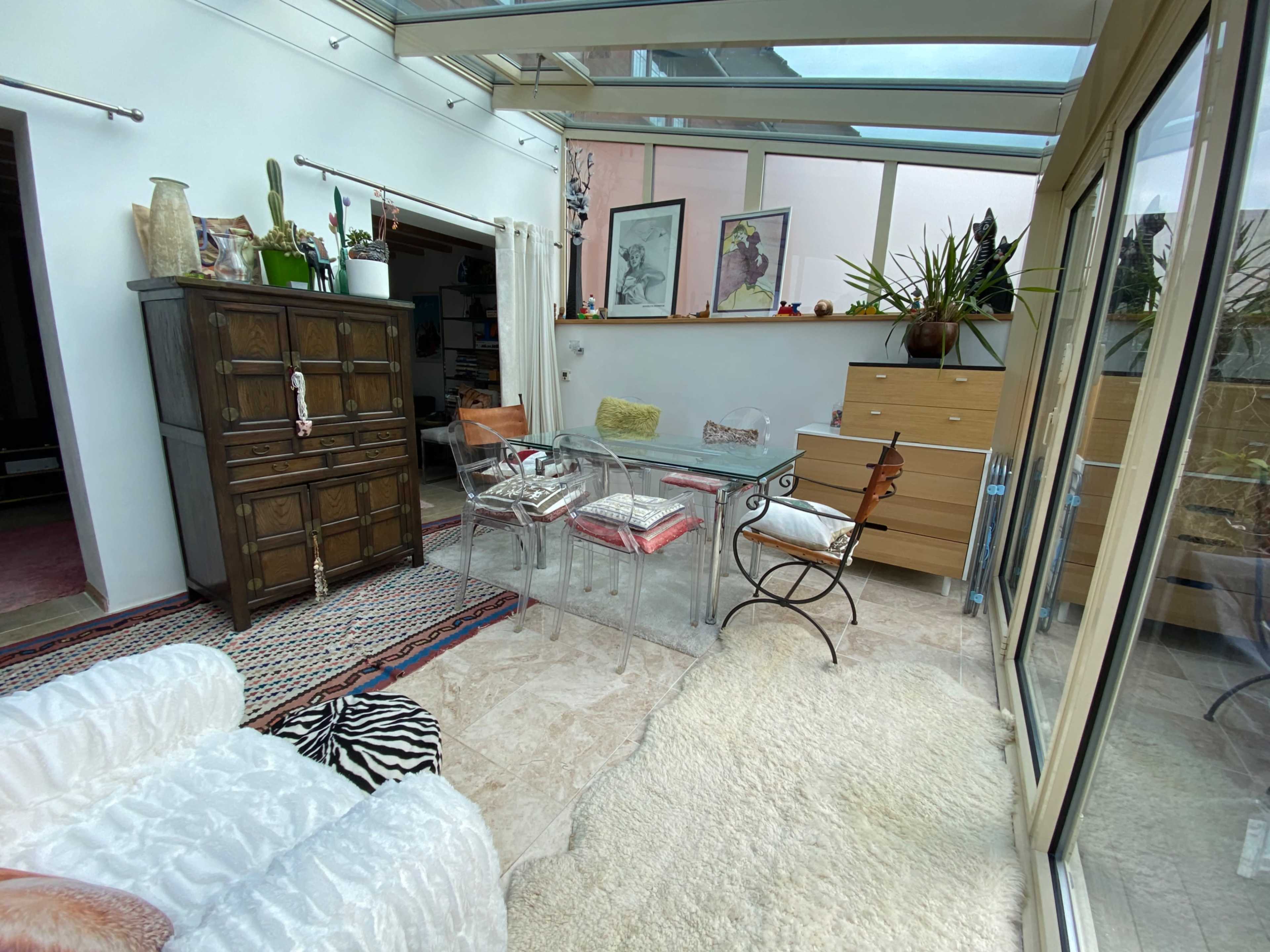 A sunlit room with a glass table surrounded by transparent chairs, a wooden cabinet, and various plants in the background.