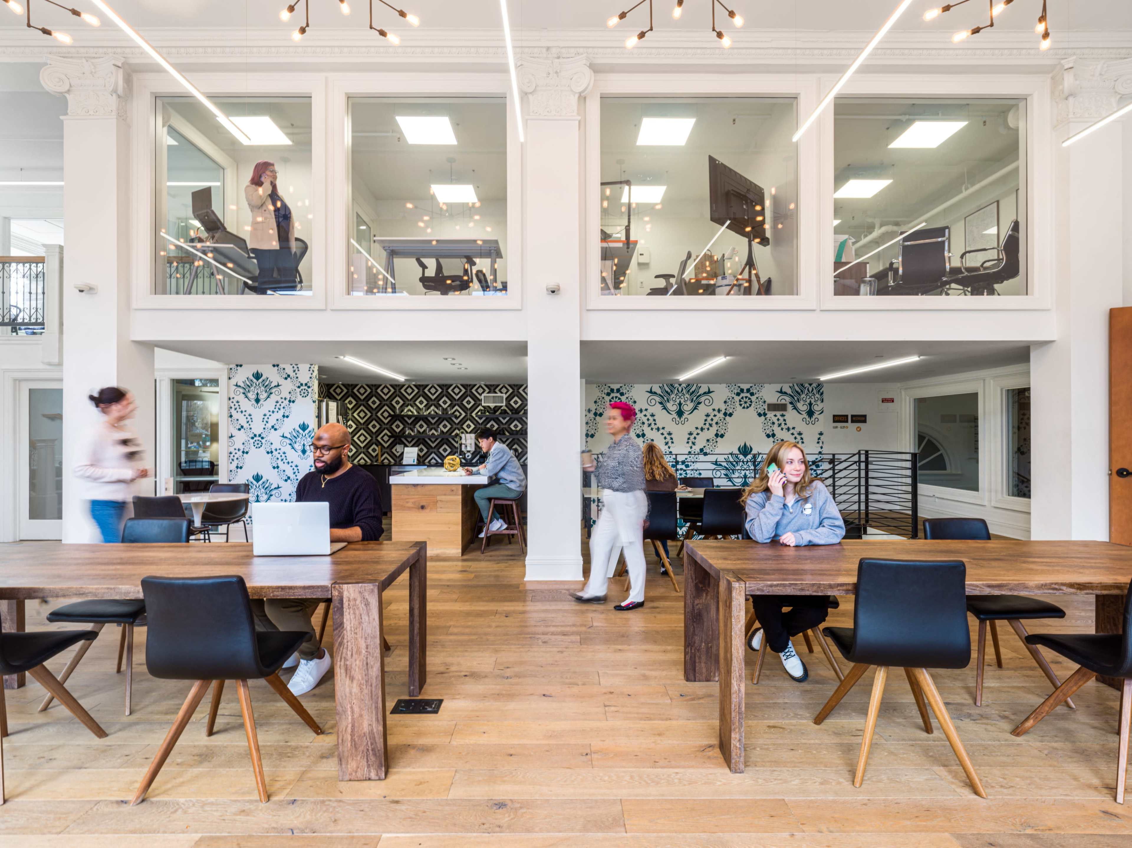 A modern co-working space features a mix of individuals working at wooden tables and others walking through an open area with large windows and decorative walls.