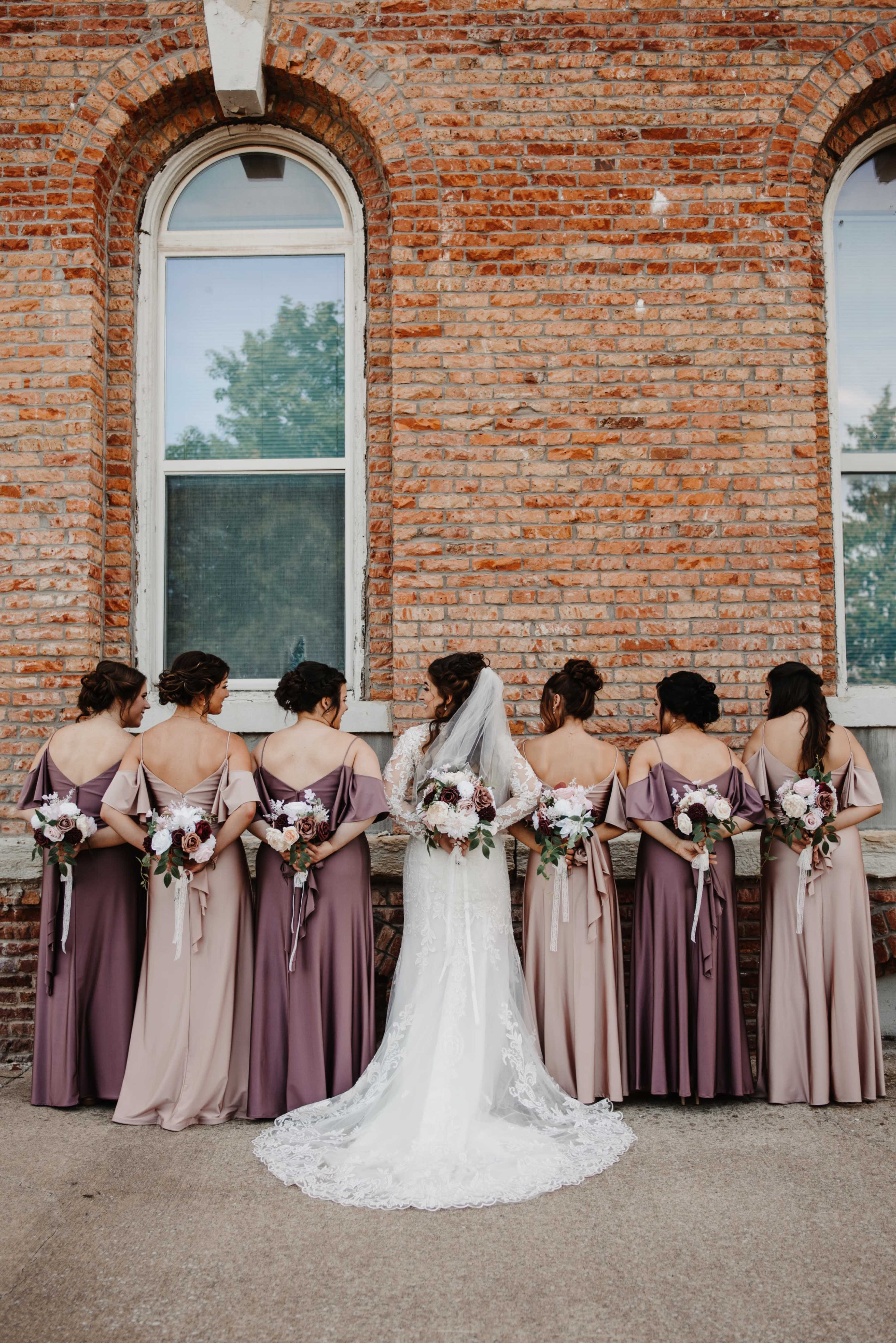 A bride in a white gown stands with her six bridesmaids, who wear long dresses in varying shades of mauve and hold bouquets, against a brick wall with large windows.