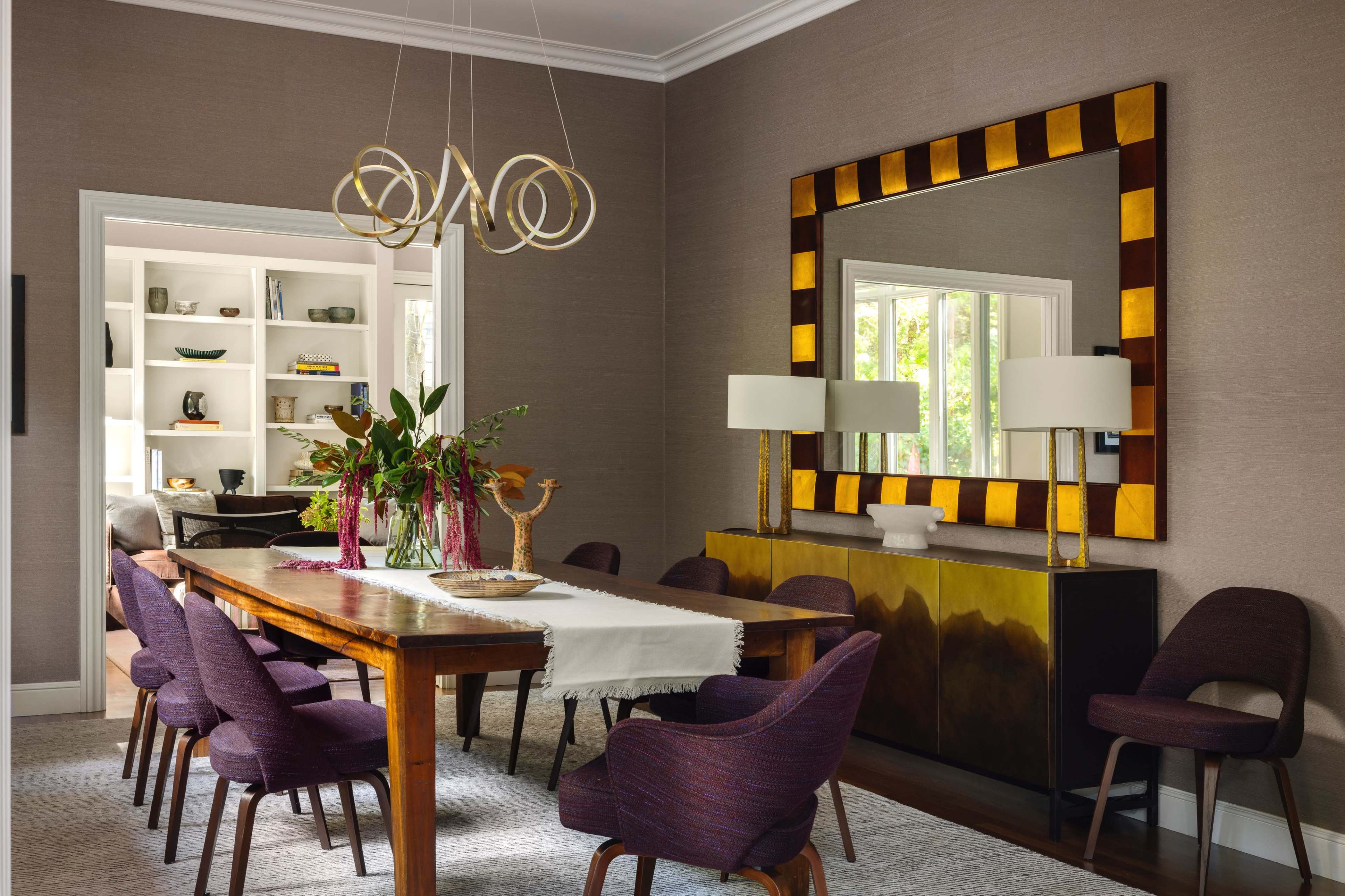 A dining room features a long wooden table surrounded by purple chairs, with a large, decorative mirror and a console table adorned with lamps and plants.