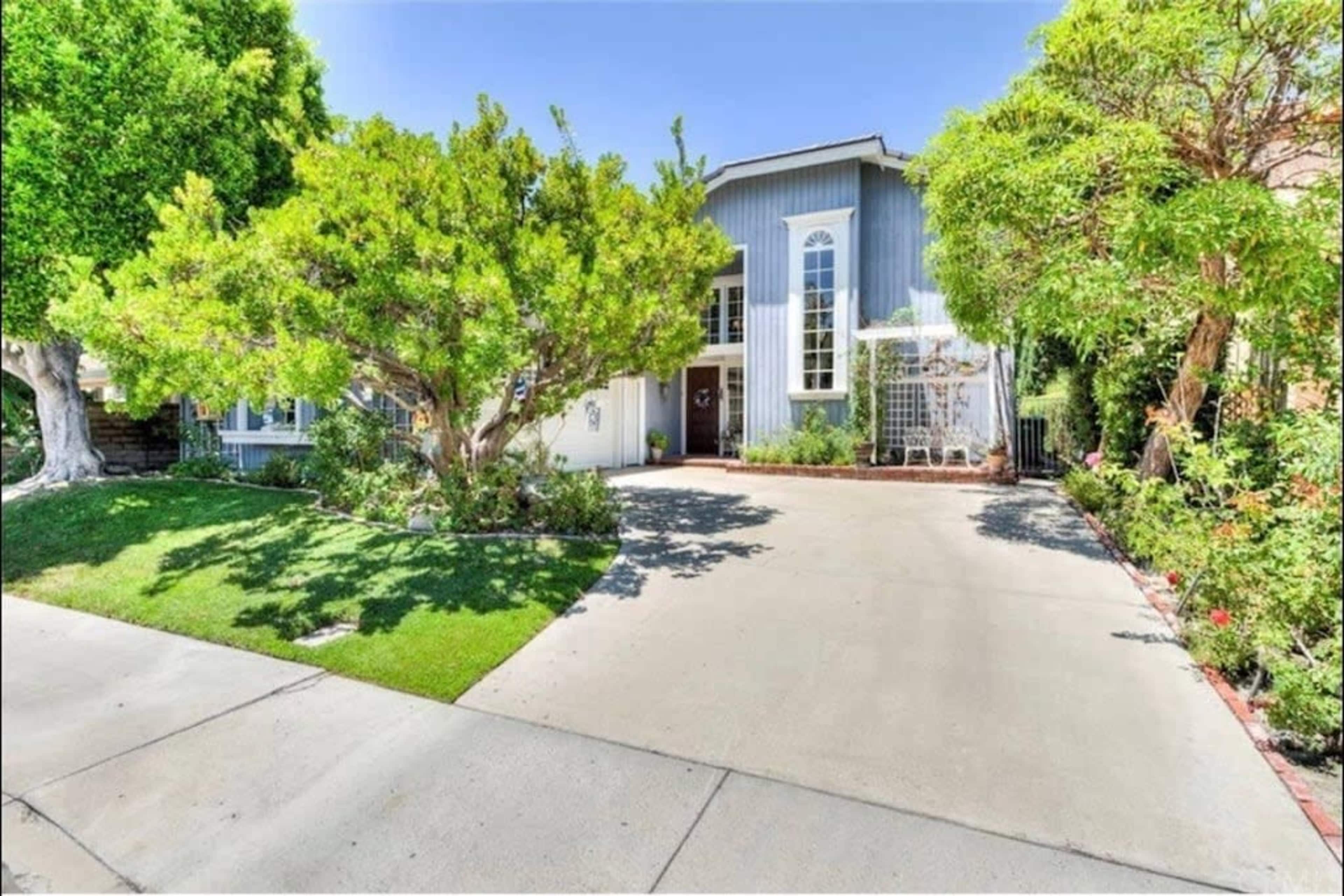 A two-story blue house with large windows is surrounded by green trees and a well-maintained lawn.