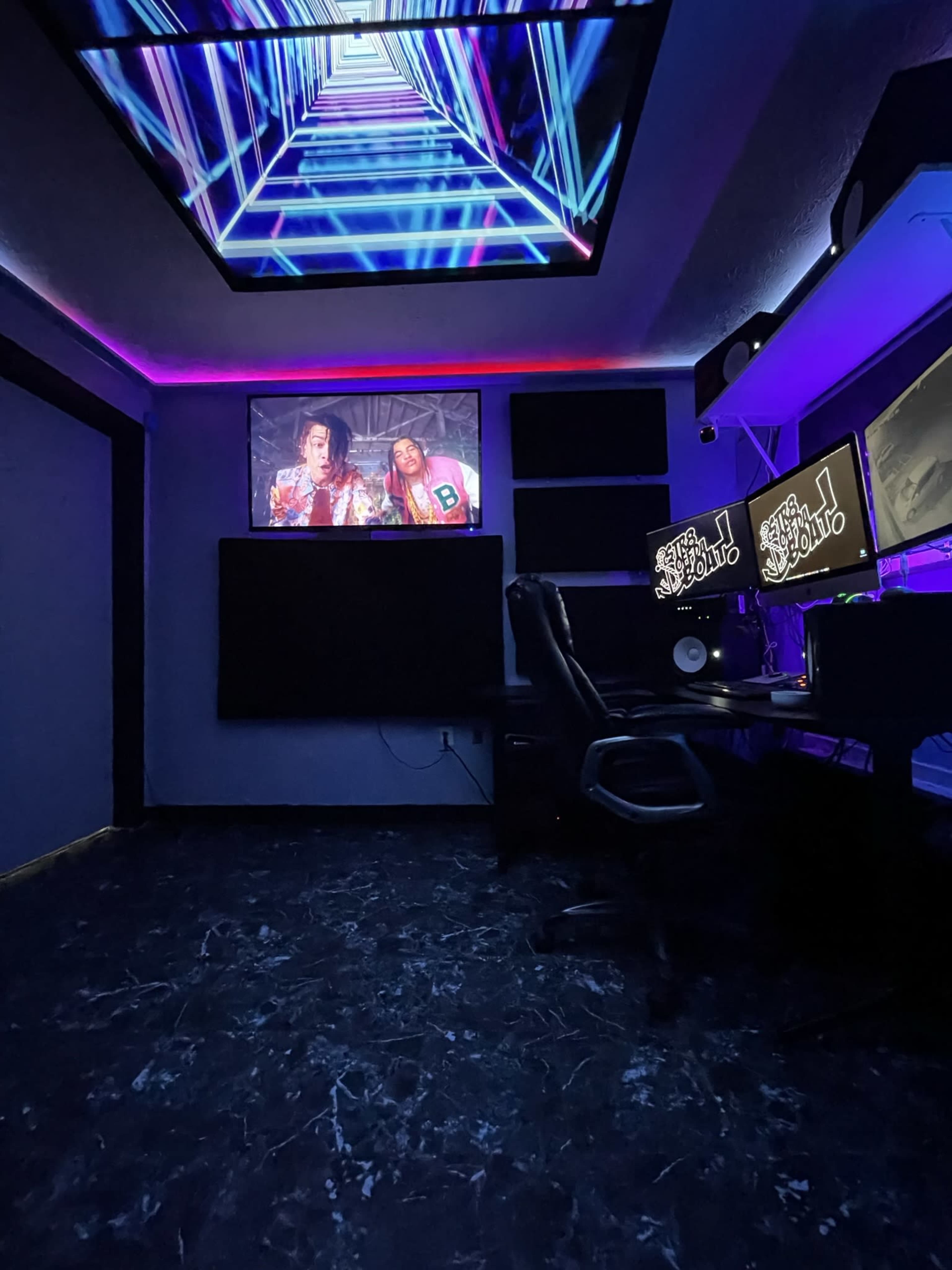 A music studio with a large screen displaying a video, neon lighting, and two computer monitors on a desk.