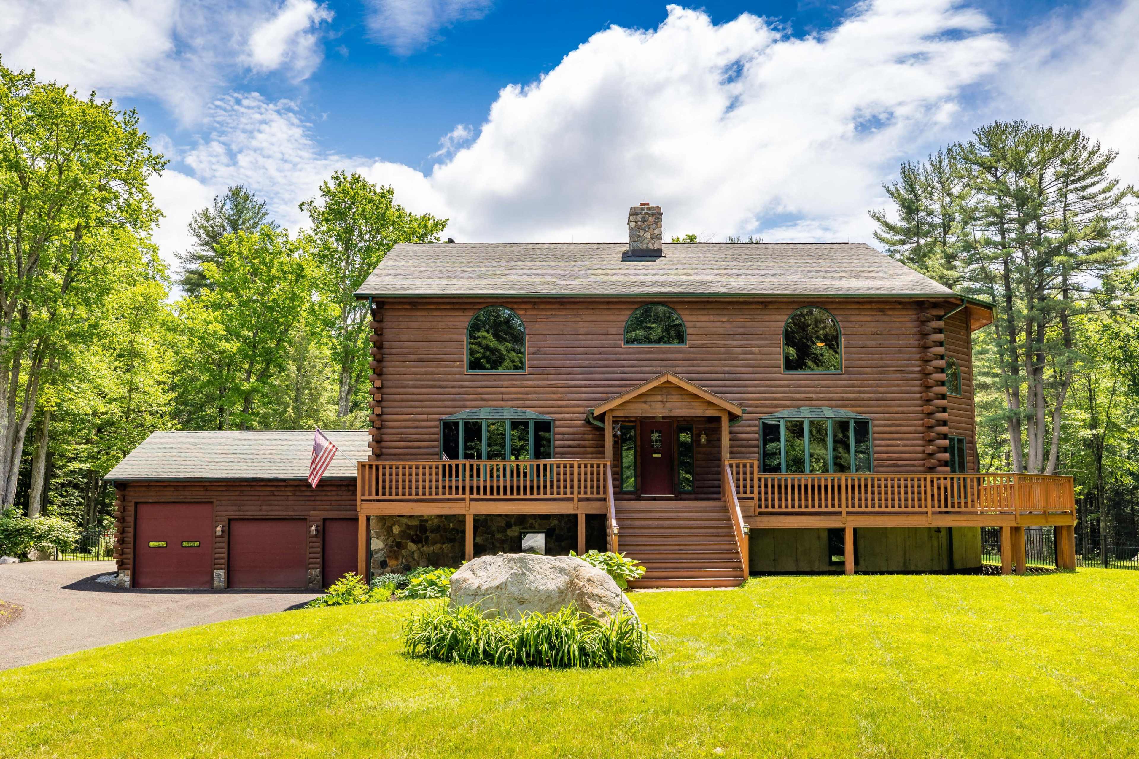 A large log cabin-style house with two garages sits on a grassy lawn surrounded by trees under a partly cloudy sky.