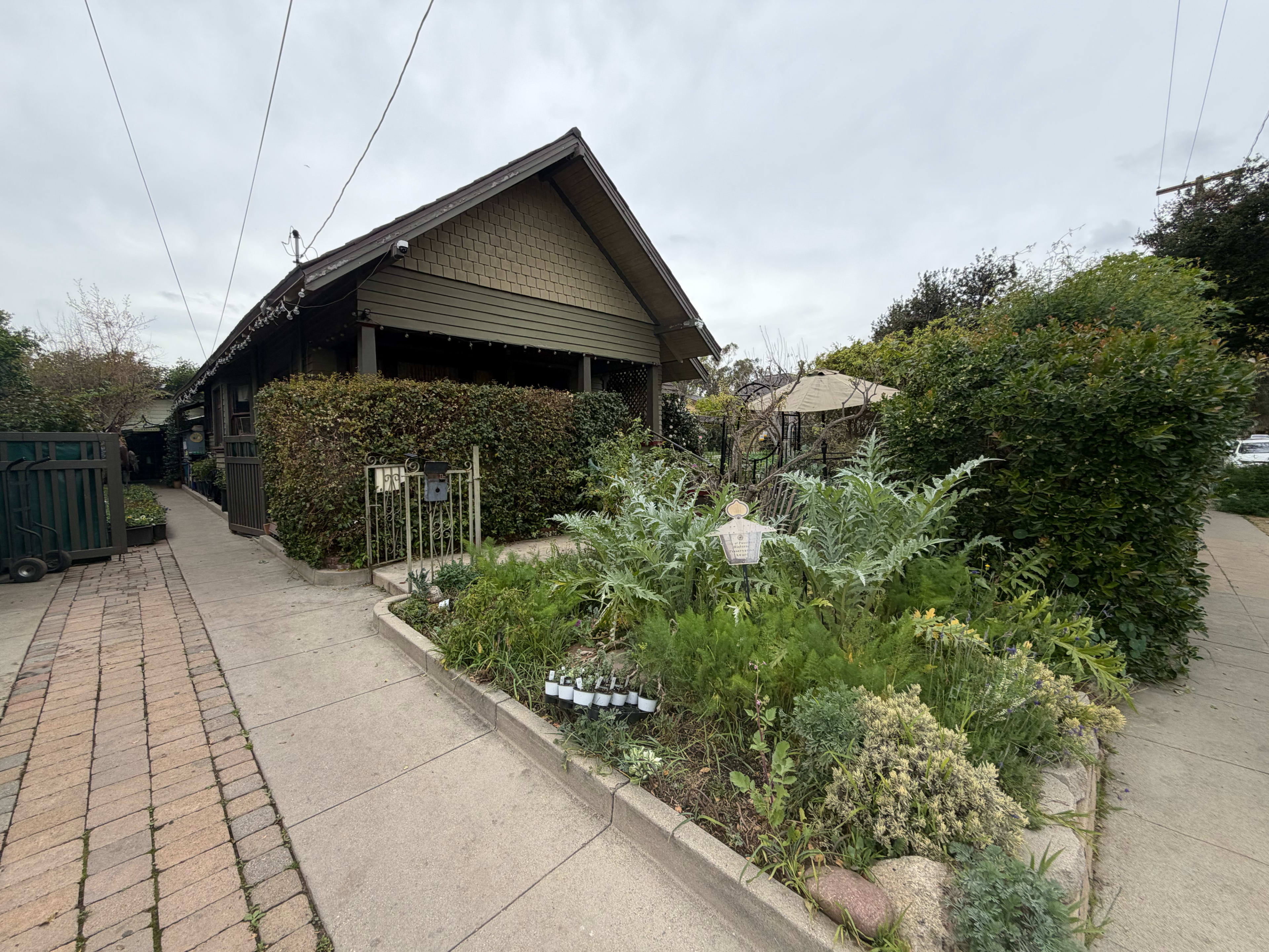 A small, green house with a well-maintained garden and a brick pathway is shown, with several plants and shrubs surrounding the entrance.
