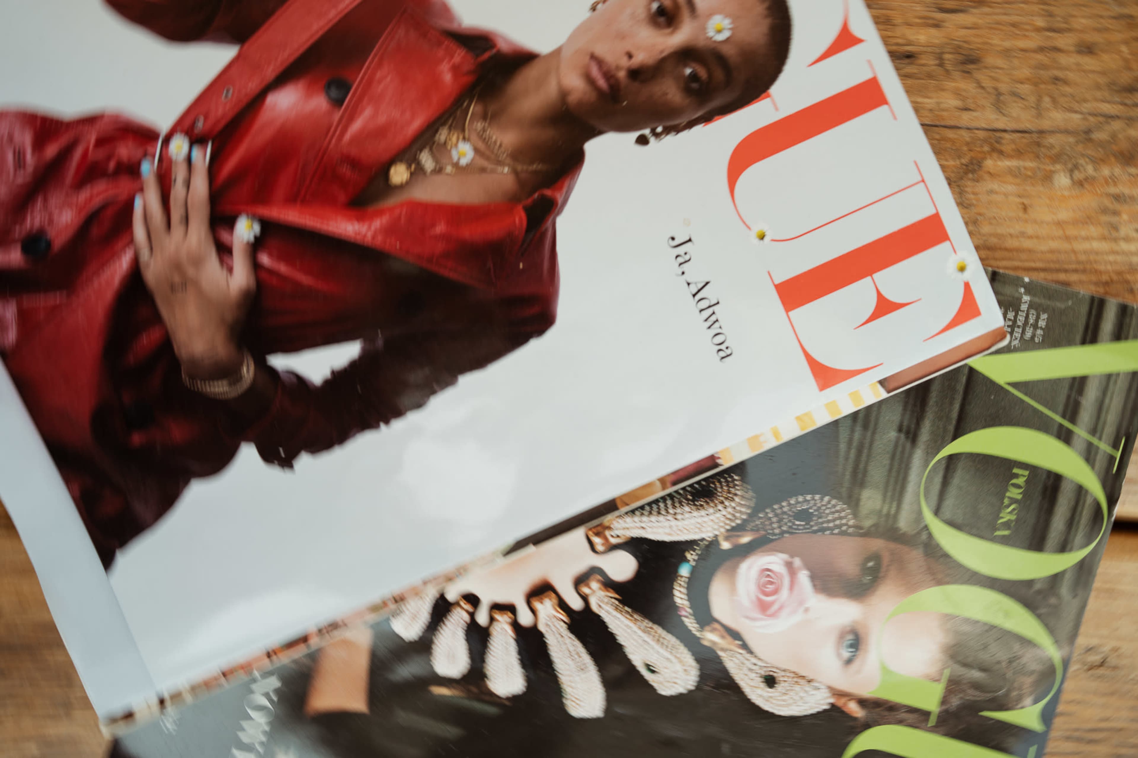 Three fashion magazines are stacked on a wooden surface, featuring bold covers with images of models in stylish outfits and jewelry.