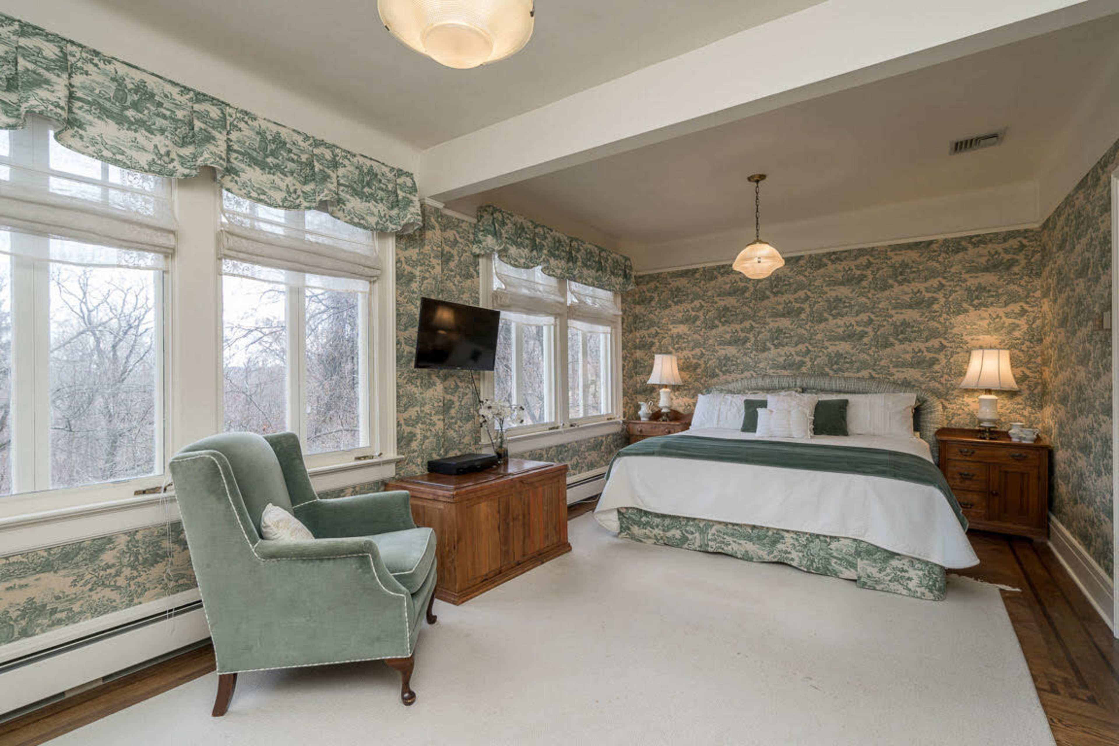 A bedroom features a bed with green and white bedding, surrounded by floral wallpaper, a light fixture, and windows that provide natural light.
