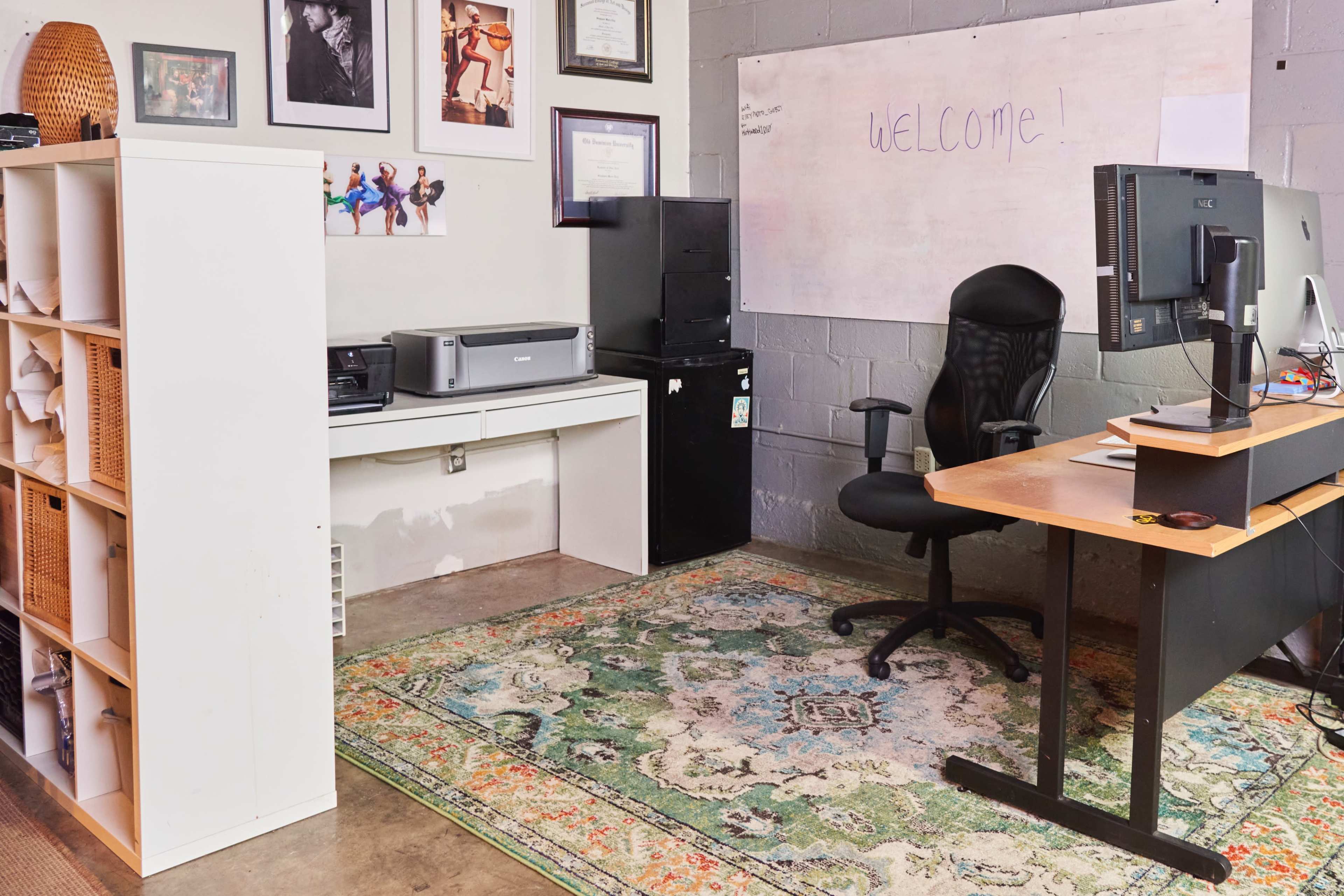 The image shows a small office space featuring a desk with a computer, a printer on a shelf, a black filing cabinet, and a rug on the floor, with a whiteboard that has "WELCOME!" written on it.