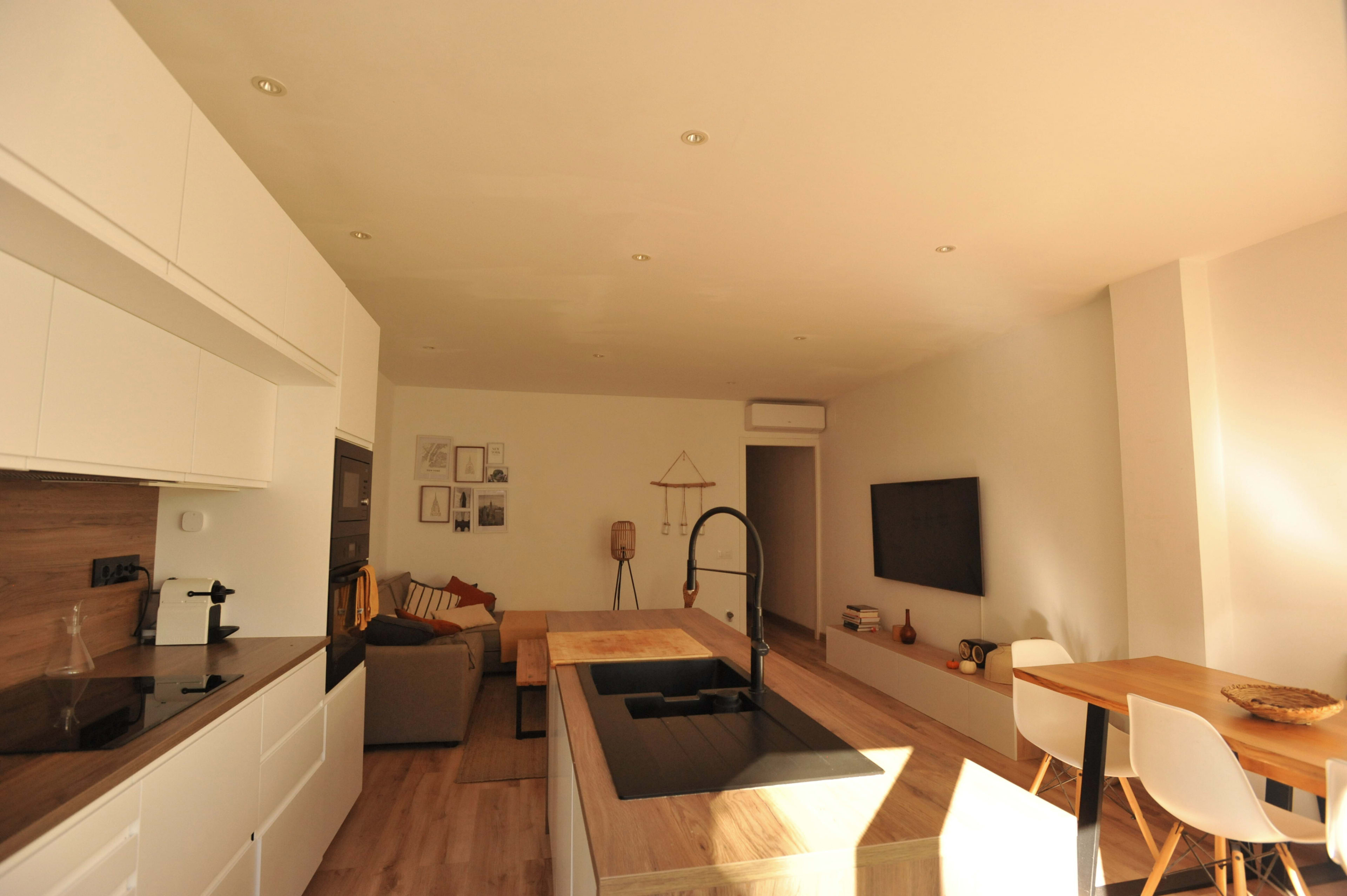 The image shows a modern, open-concept kitchen and living area with a central island, dining table, seating area, and wall-mounted television.