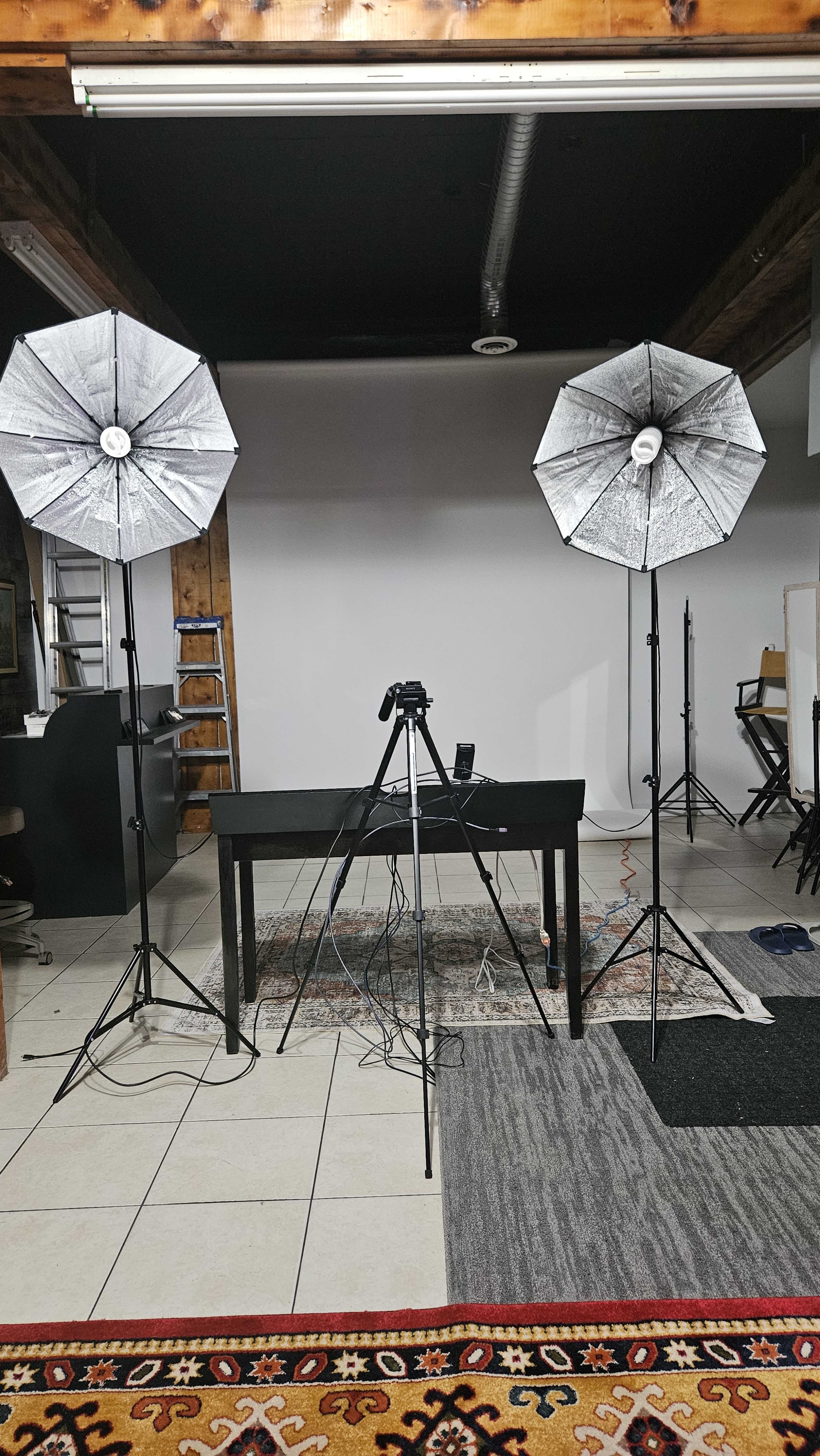 The image shows a studio setup with two softbox lights, a camera on a tripod, and a table in front of a plain backdrop.