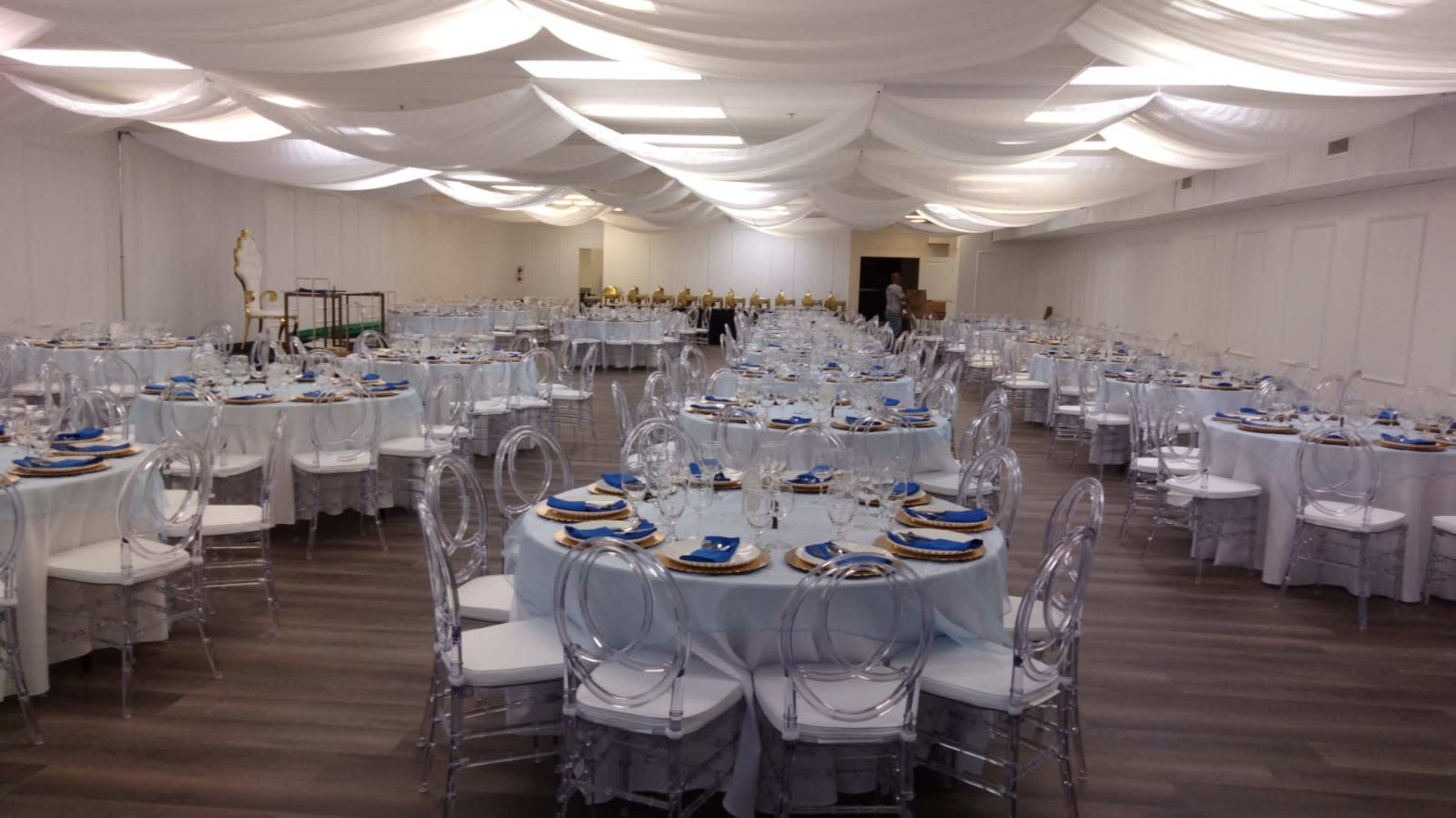 The image shows a large banquet hall set up with multiple round tables, each adorned with light blue tablecloths, white chairs, and elegant place settings.