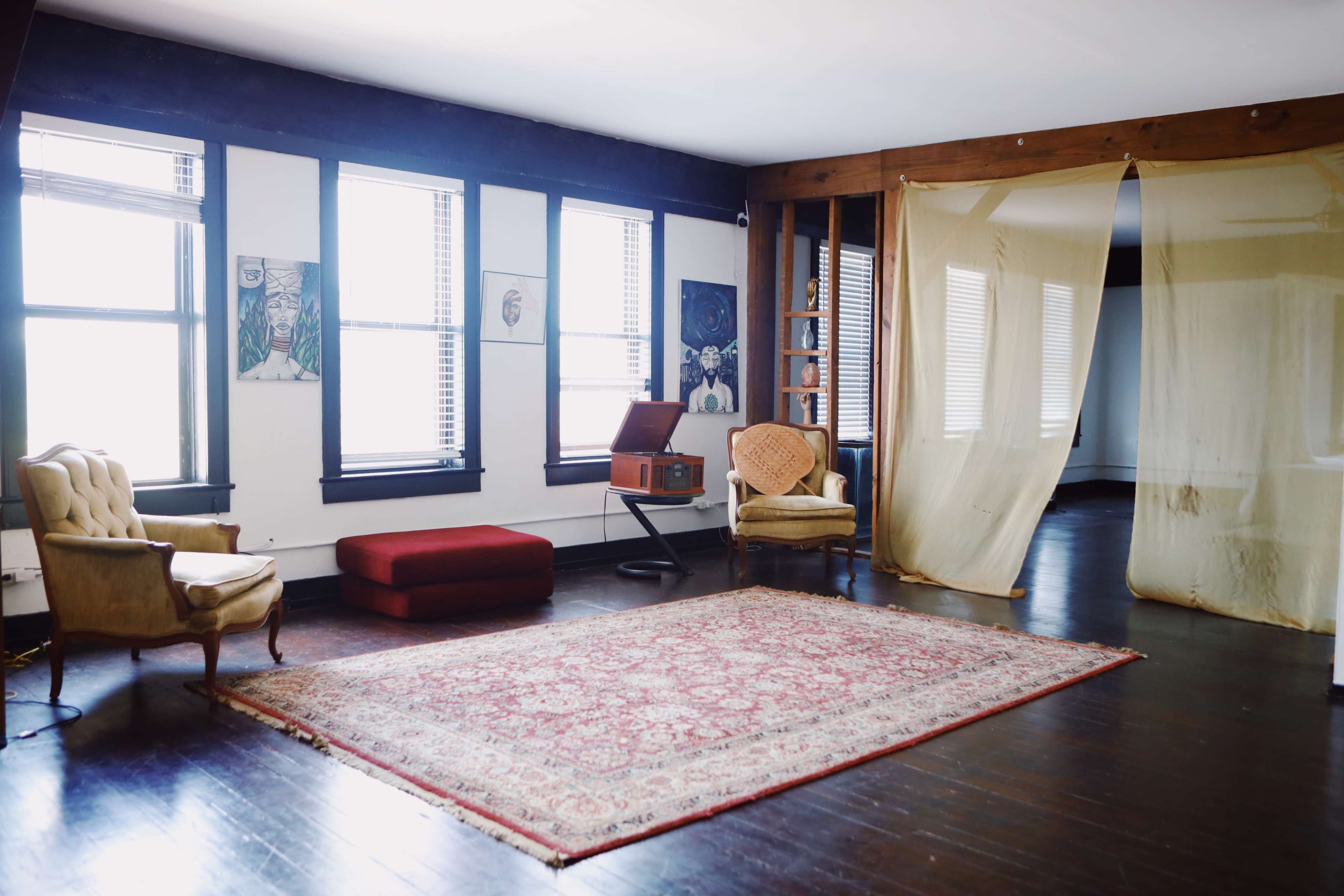 A spacious room with large windows, a patterned rug on the floor, a vintage record player, an armchair, and a curtain partitioning a side area.