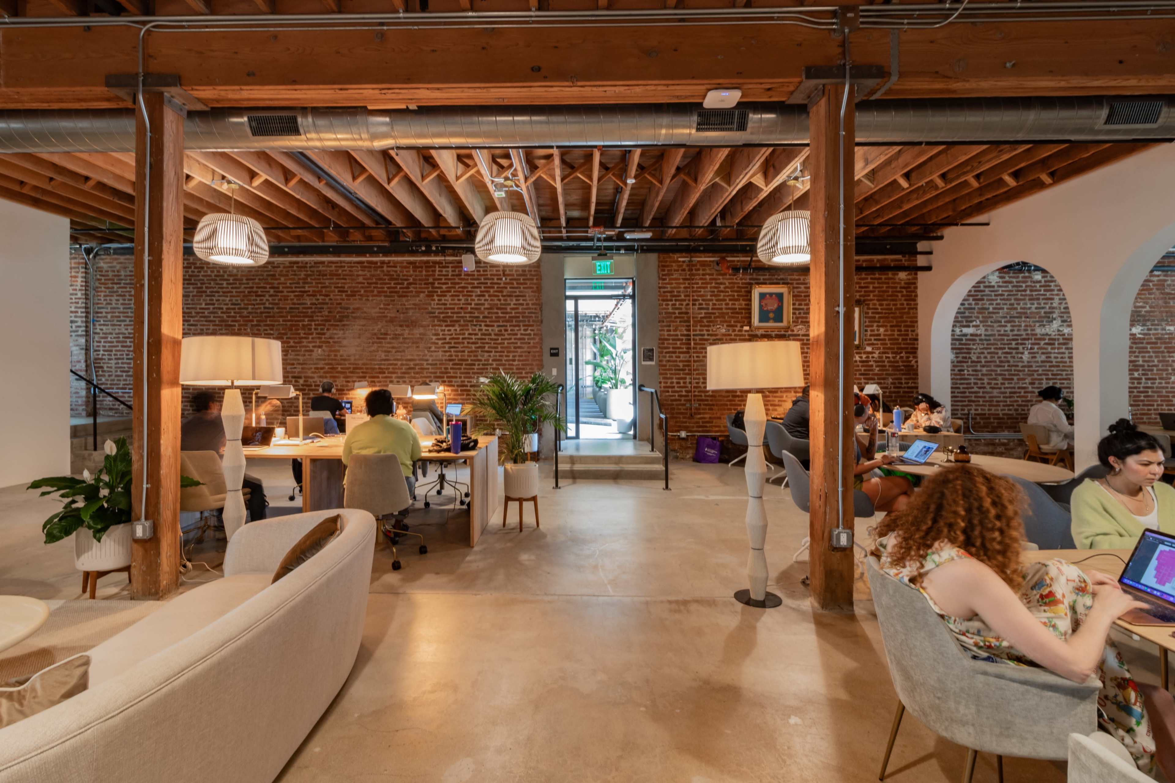 The scene shows a spacious coworking area with several people working at desks, surrounded by exposed brick walls and wooden beams.