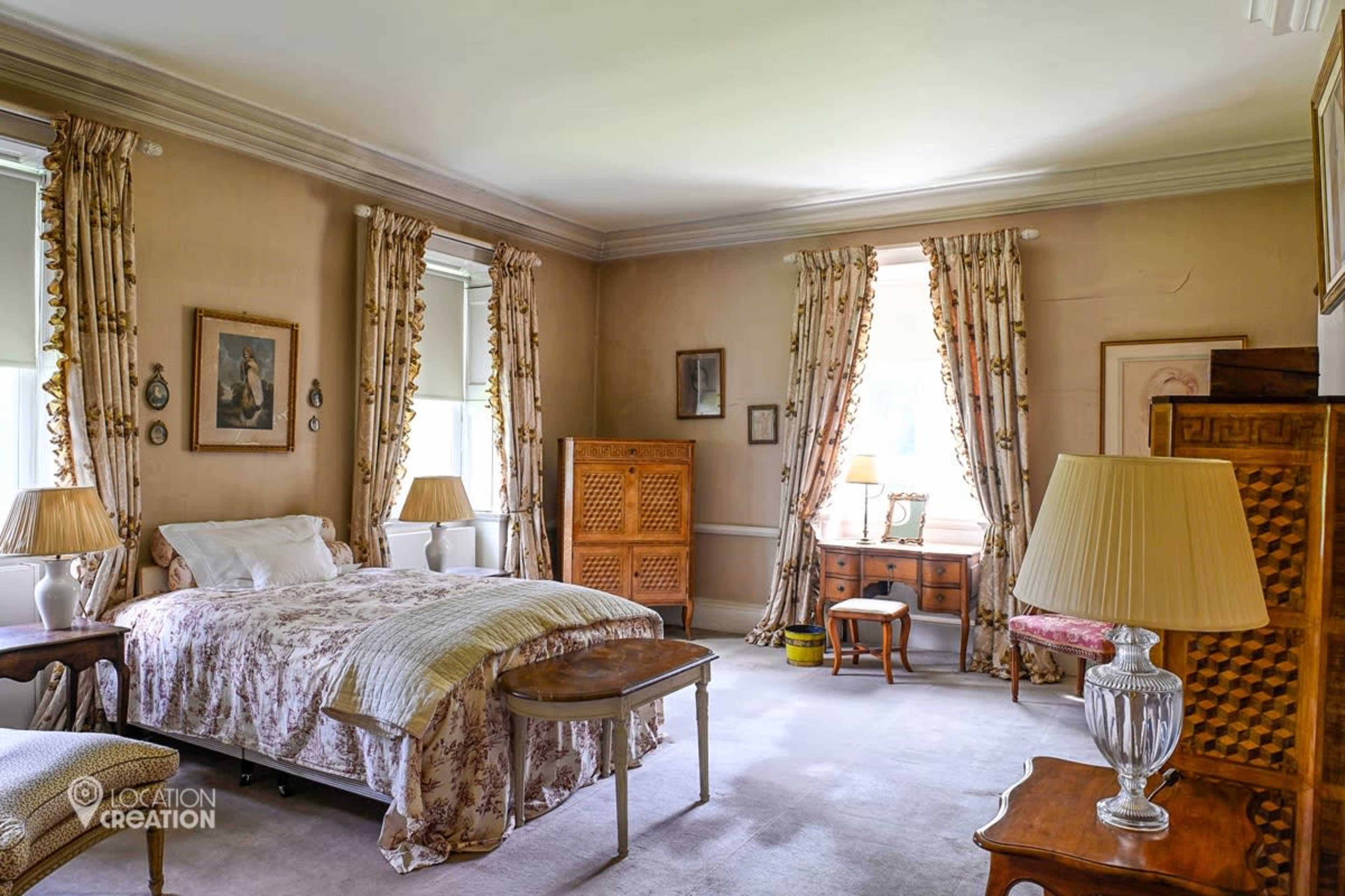 The room features a bed with floral bedding, wooden furniture, and large windows dressed with patterned curtains.