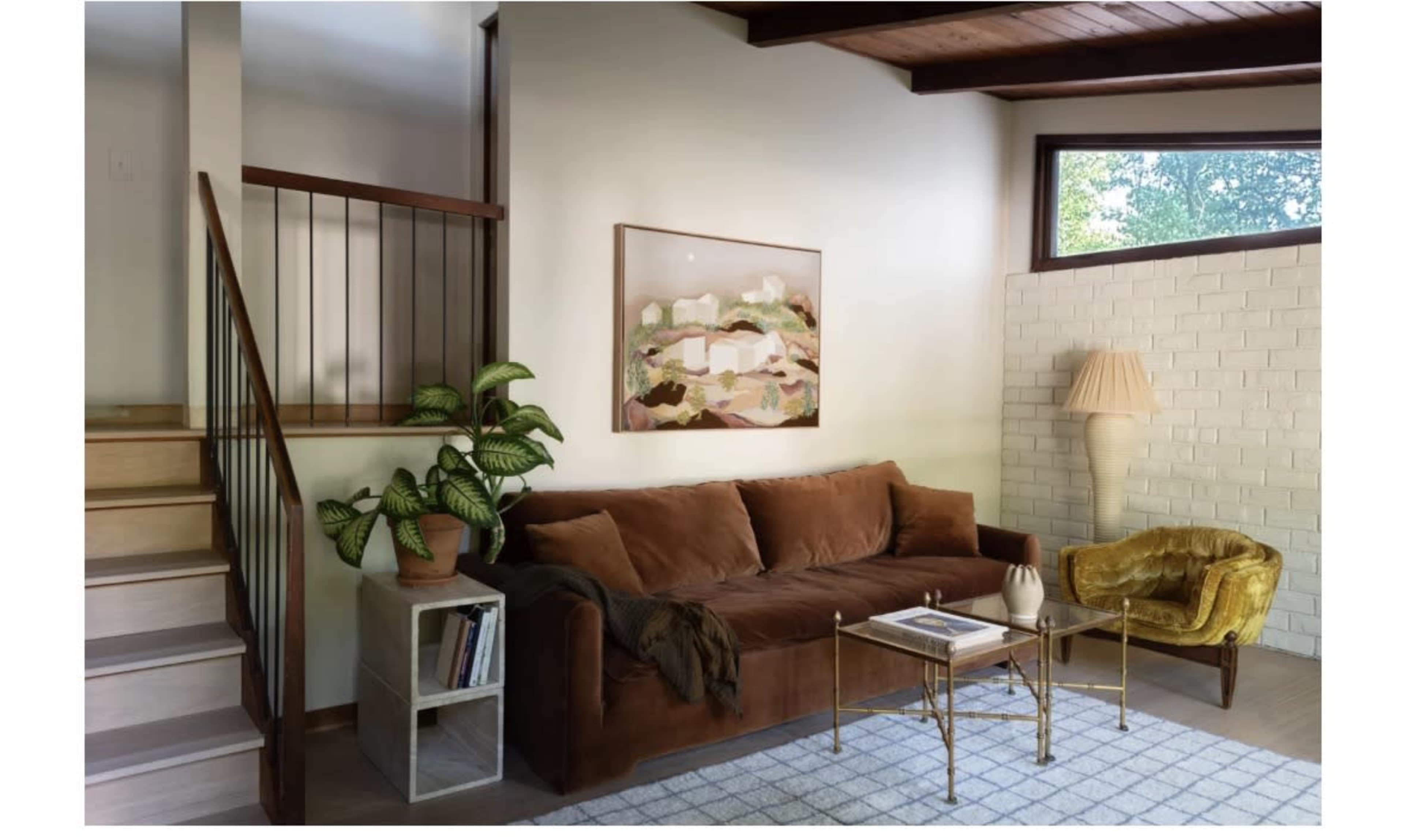 The image shows a modern living room featuring a brown sofa, a glass coffee table, a yellow accent chair, and a staircase in the background.