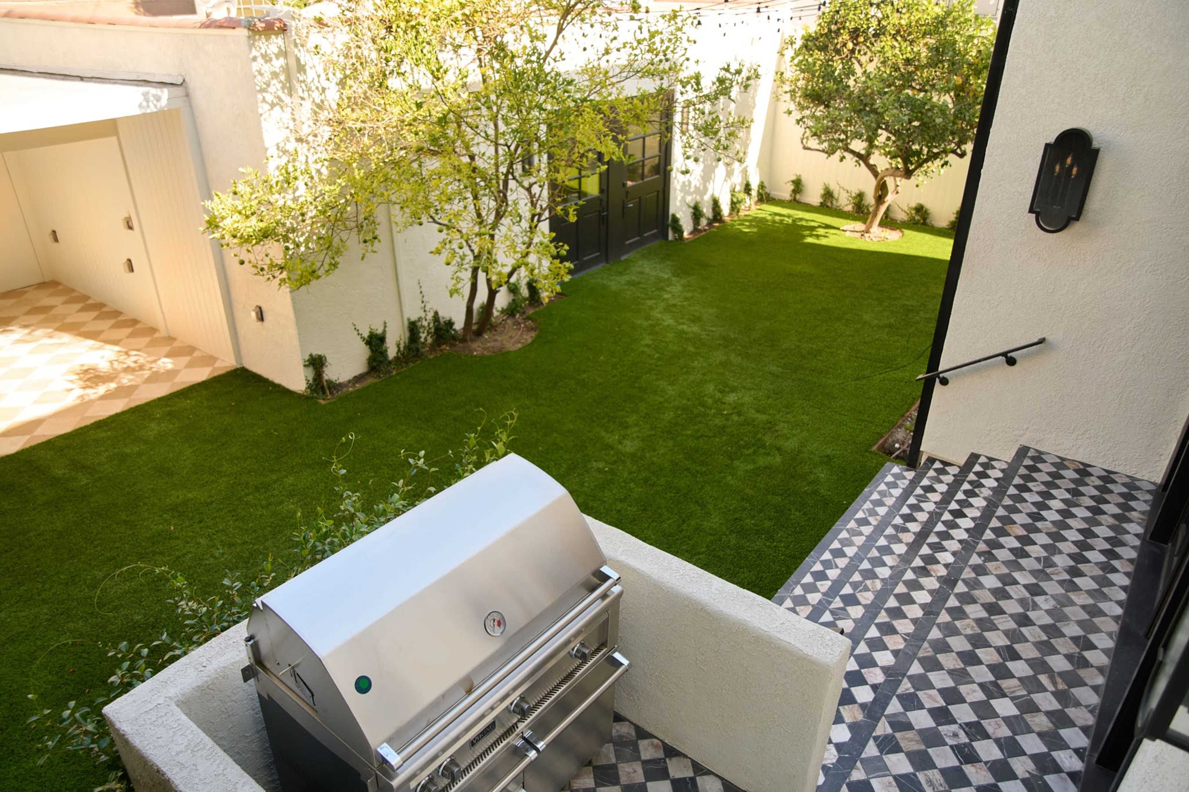 The image shows a well-manicured backyard with a green lawn, a grill on a patio, and trees surrounding a black door leading to a structure.