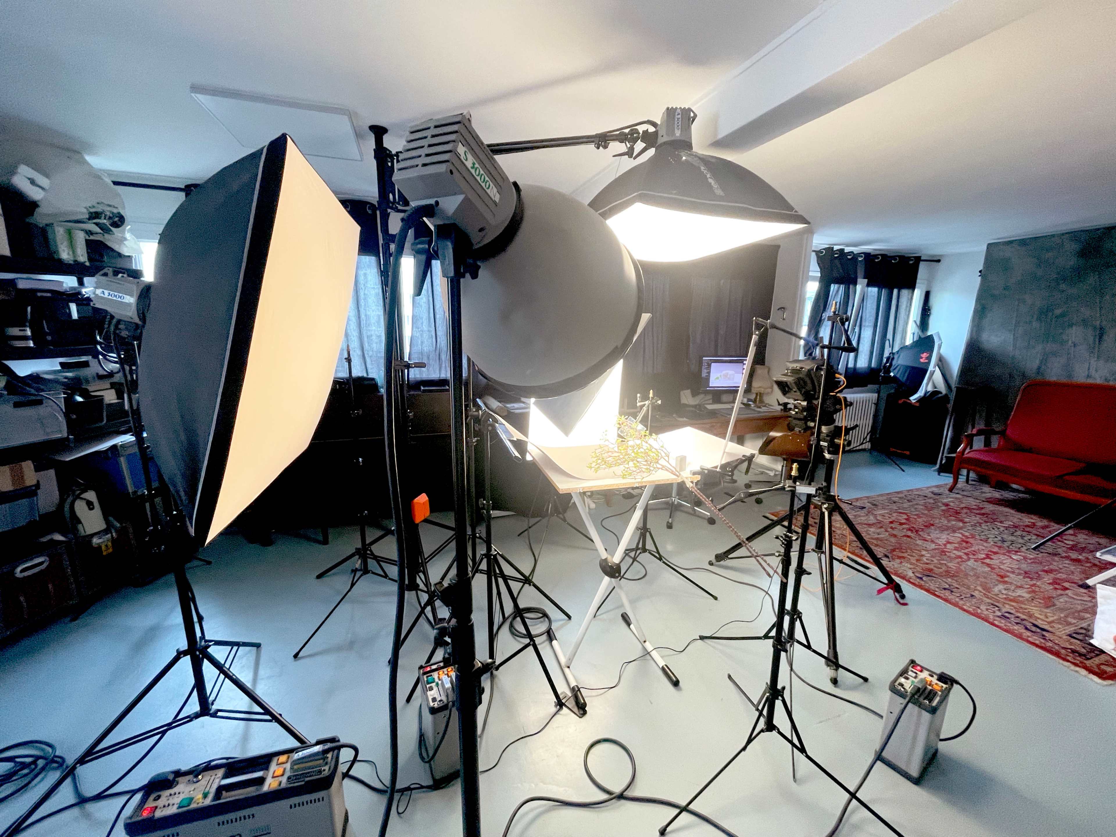 The image shows a photography studio setup with multiple light sources, a table arranged with equipment, and a red sofa in the background.