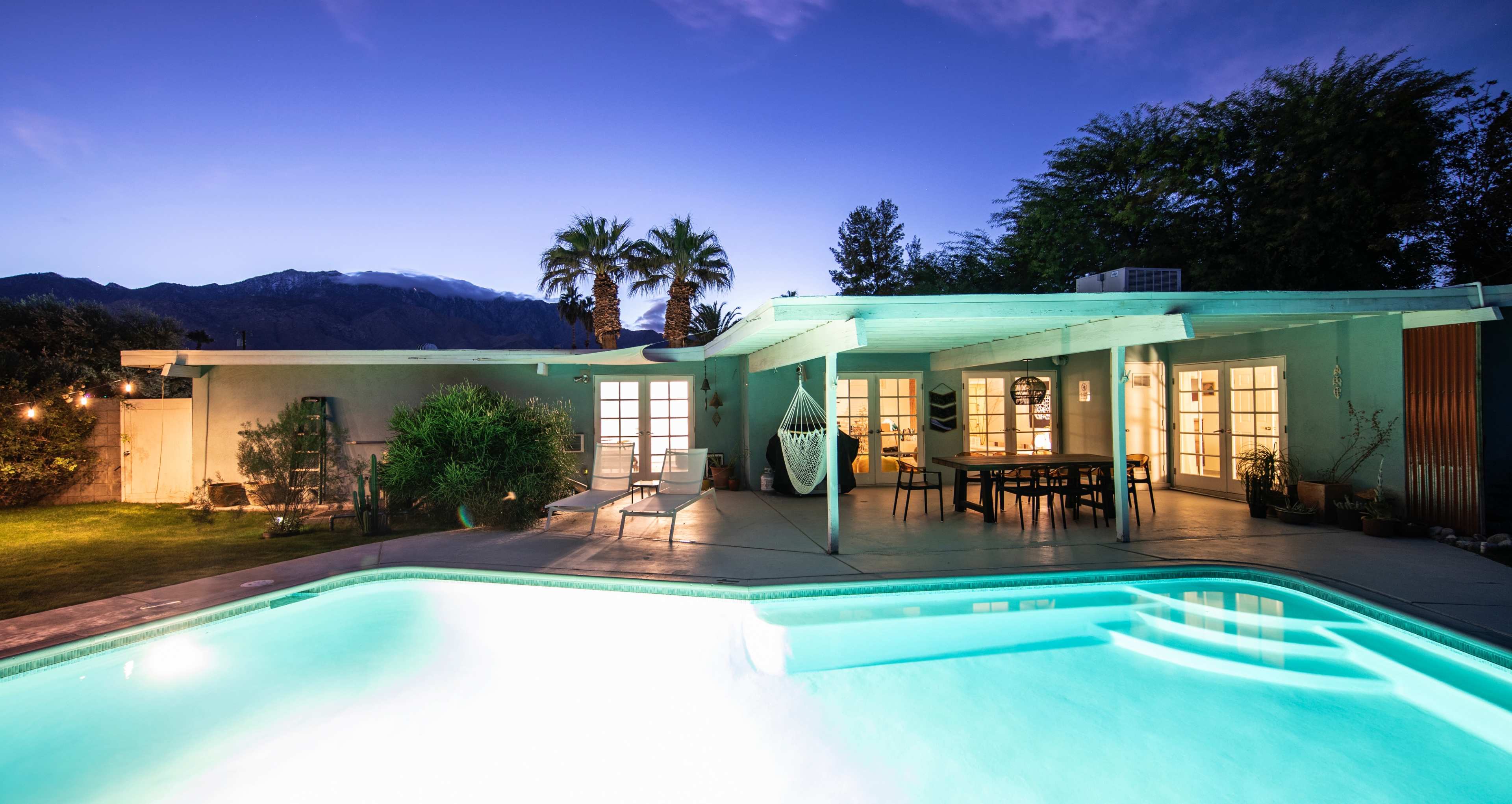 A poolside view of a modern home at dusk, with a clear sky and mountains in the background.