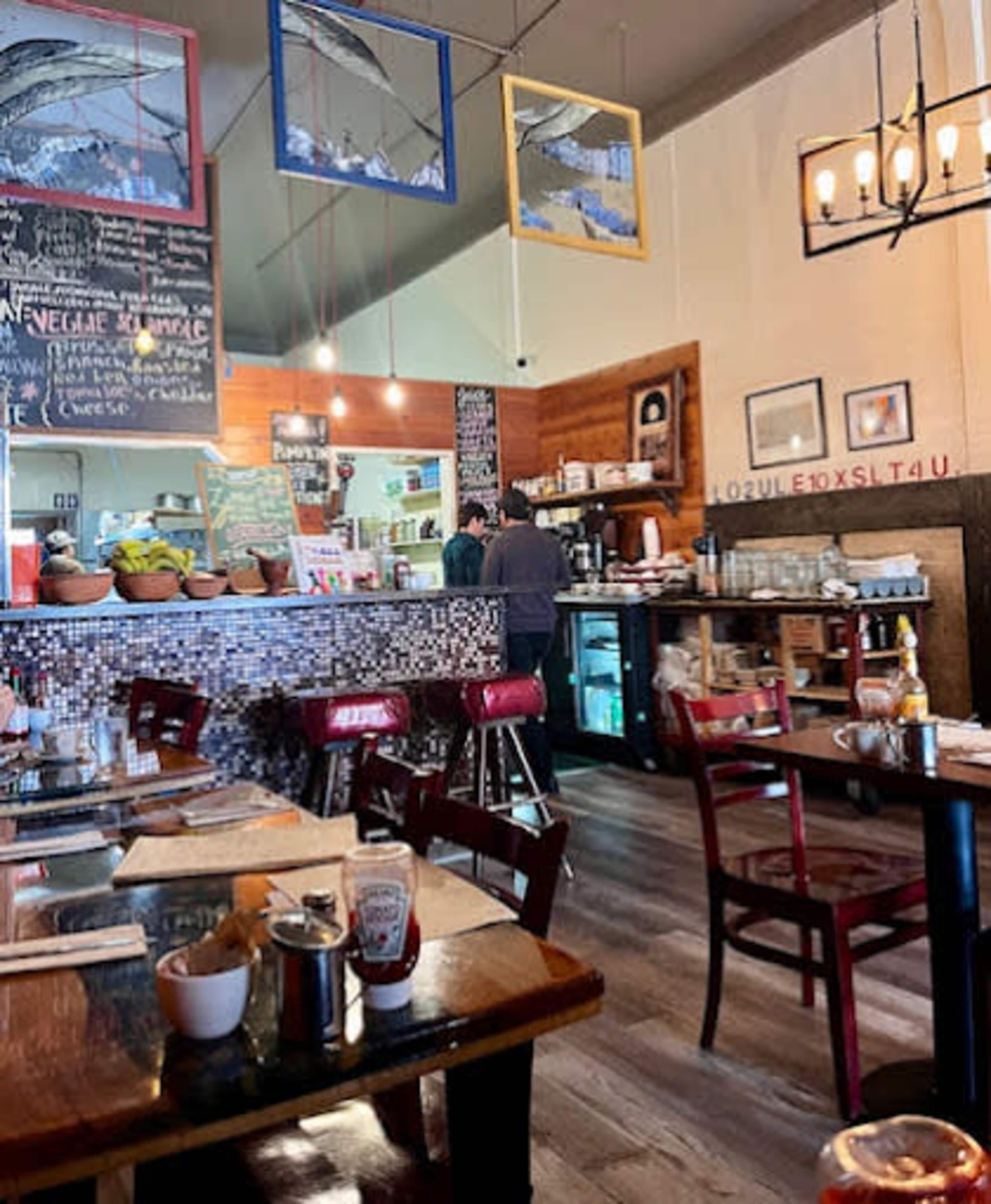 The image shows the interior of a restaurant with wooden tables, red chairs, a bar area with various food items, and chalkboard menus on the walls.