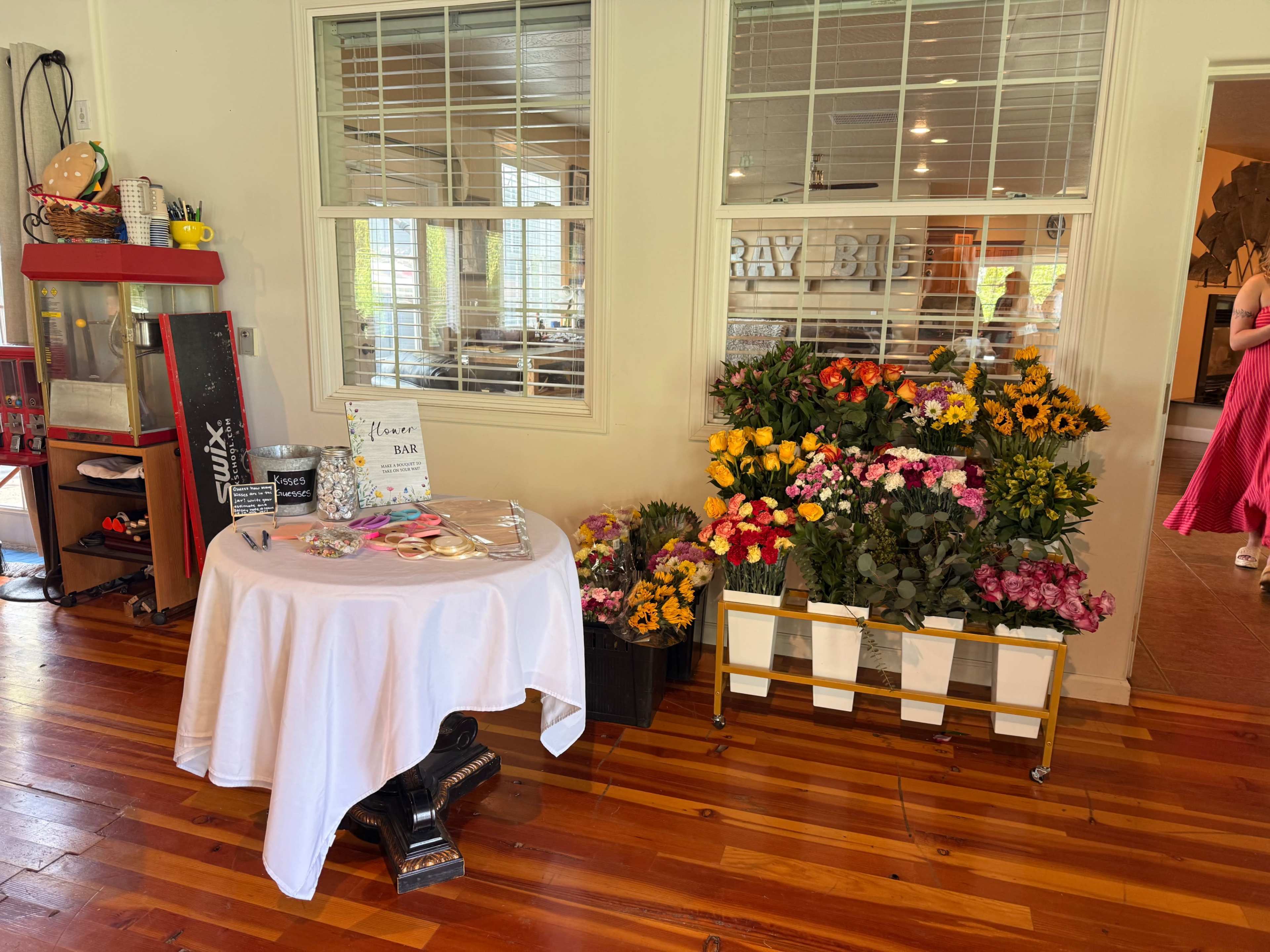 The image shows a table draped in white fabric with cookies on display, surrounded by vibrant flower arrangements in crates against a wooden floor in a brightly lit room.