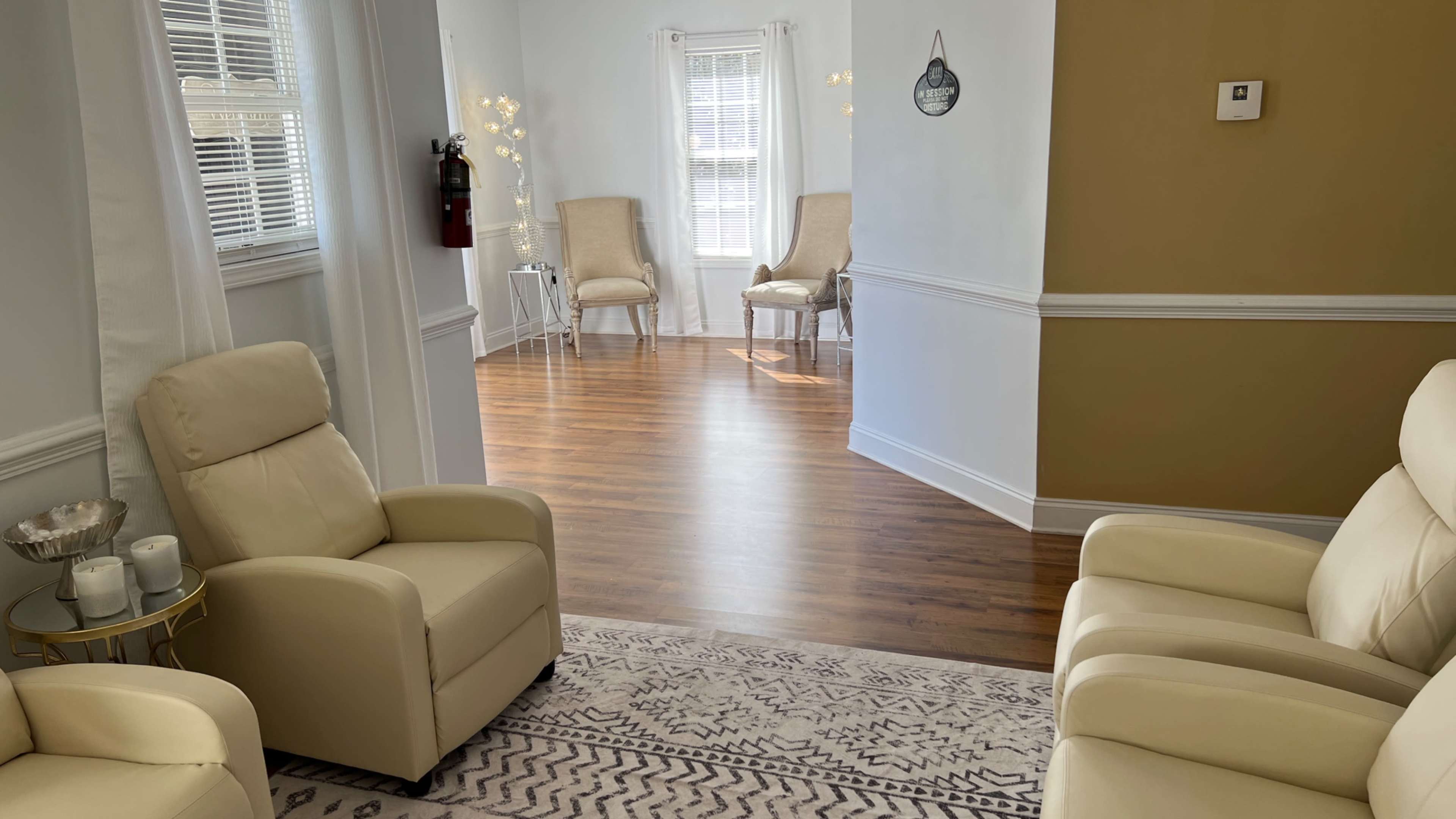 The image shows a well-lit room with two beige chairs facing a doorway leading to another area with wooden flooring, light decor, and two additional chairs.