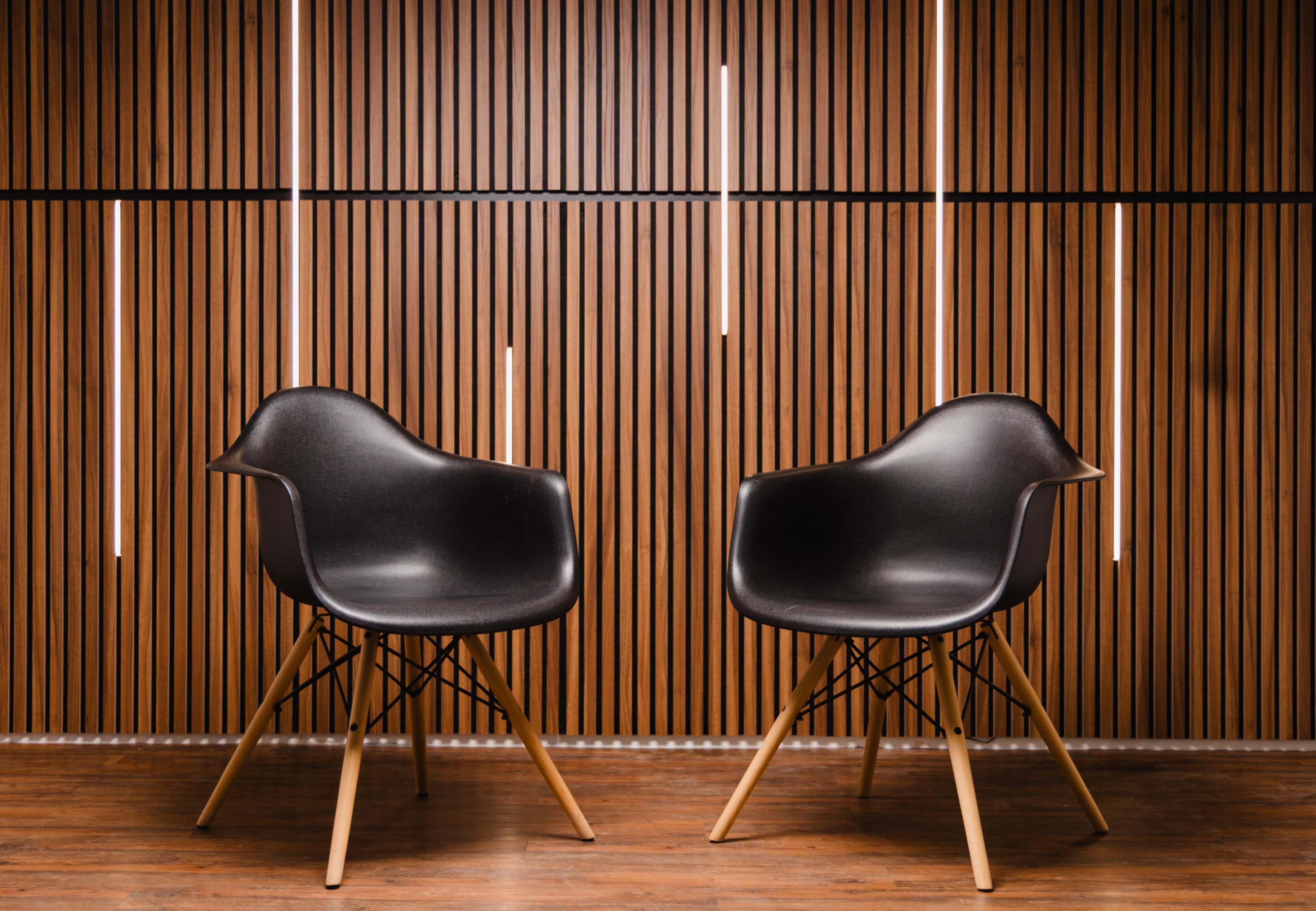 Two black molded chairs with wooden legs are positioned in front of a wooden slat wall illuminated by vertical light fixtures.
