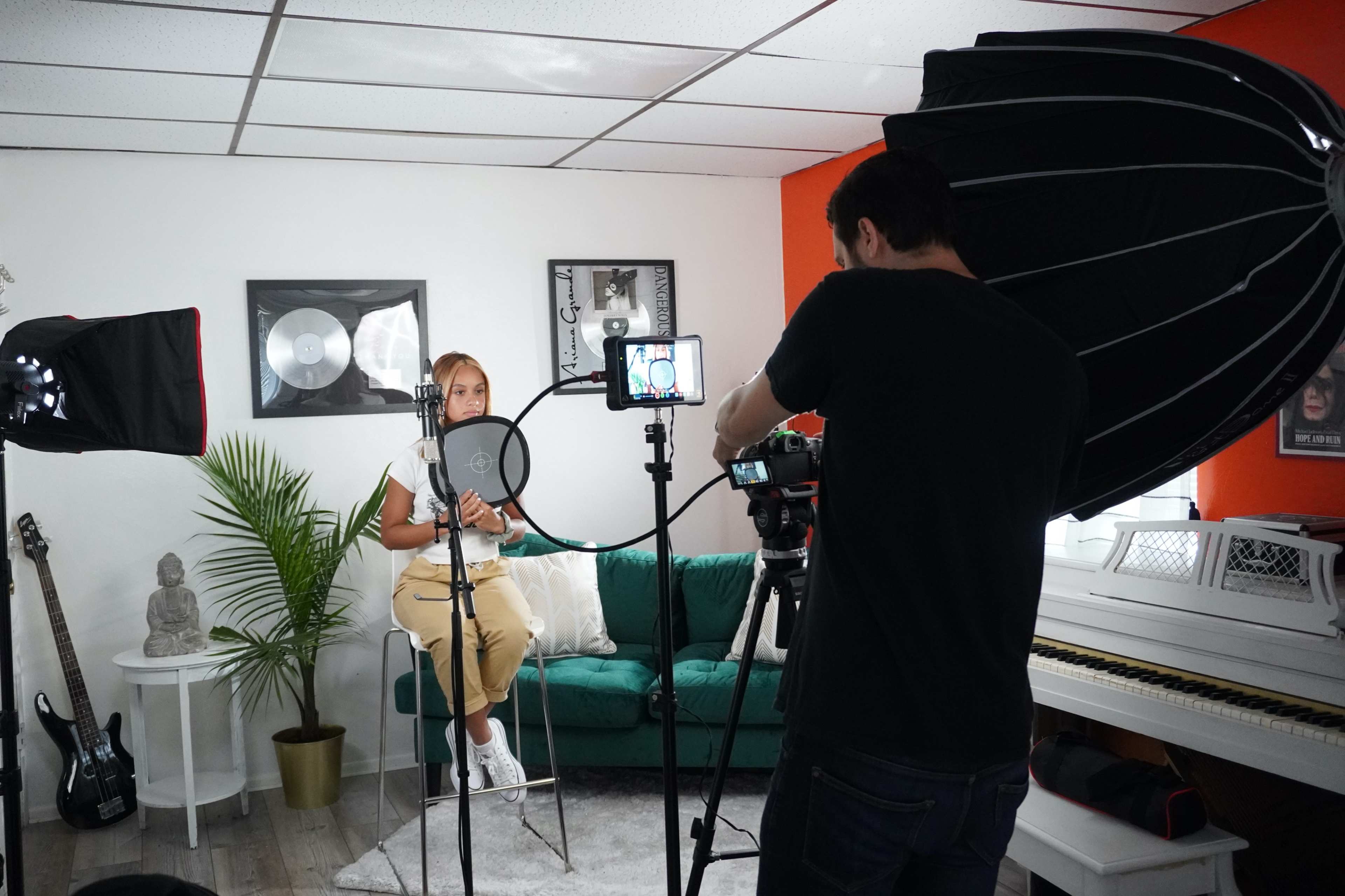 A woman sits on a teal couch in a studio setting while a man operates a camera with bright lights positioned around them.