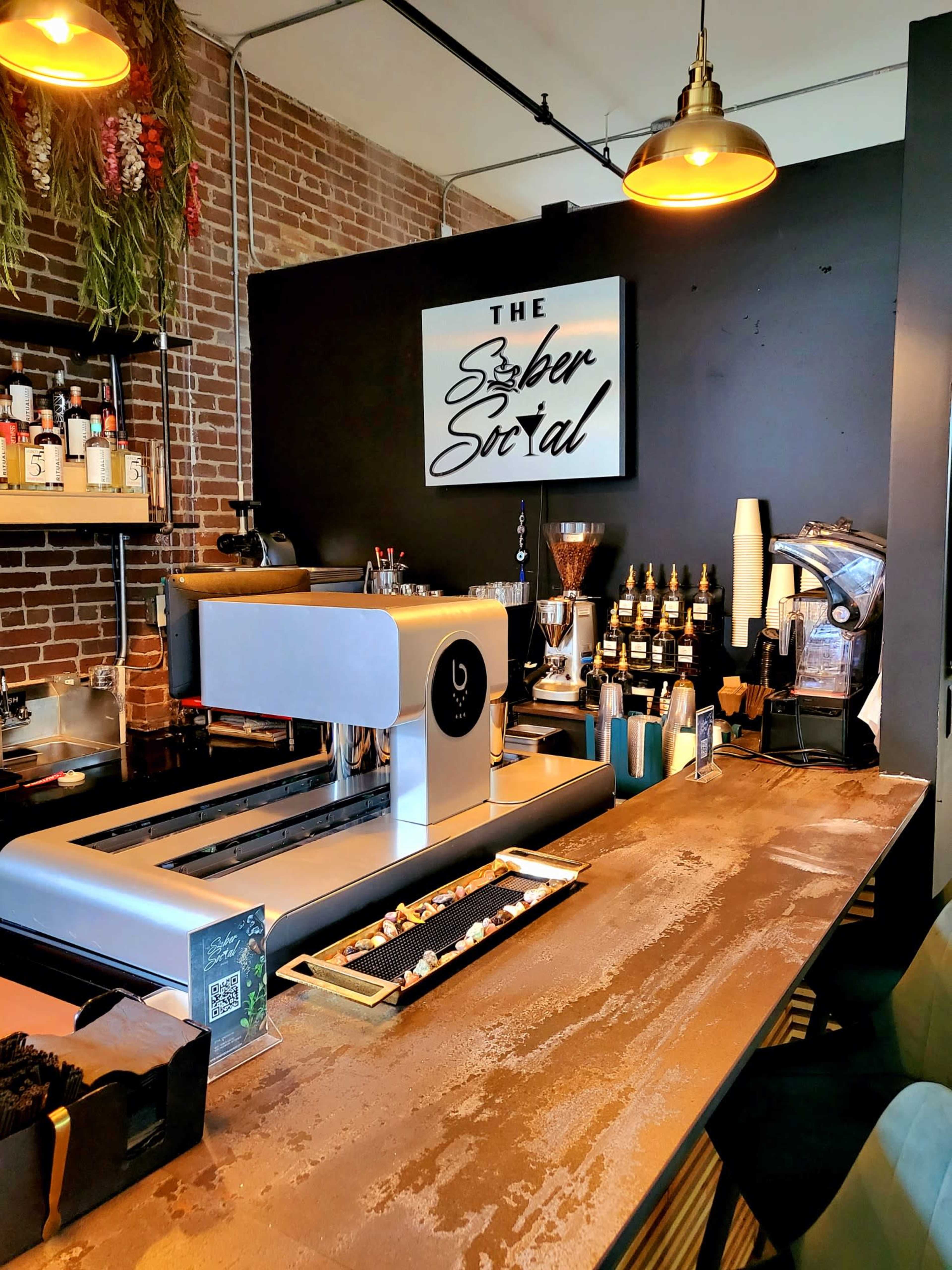 A modern coffee bar with a sleek espresso machine, a variety of bottles and equipment on display, and a dark brick wall adorned with decorative plants.