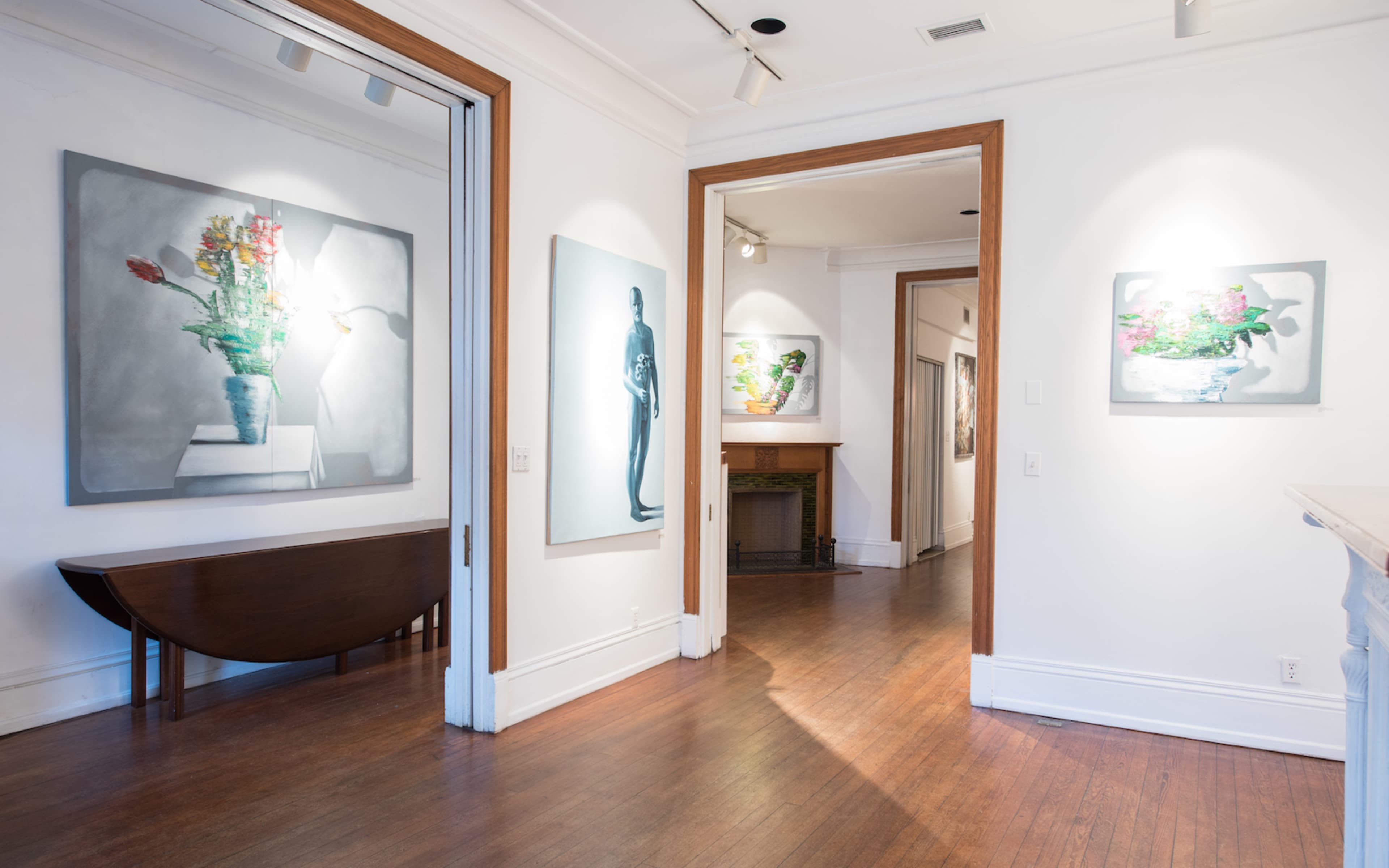 A gallery interior with artwork displayed on white walls, including floral and figure paintings, and a wooden table in the foreground.