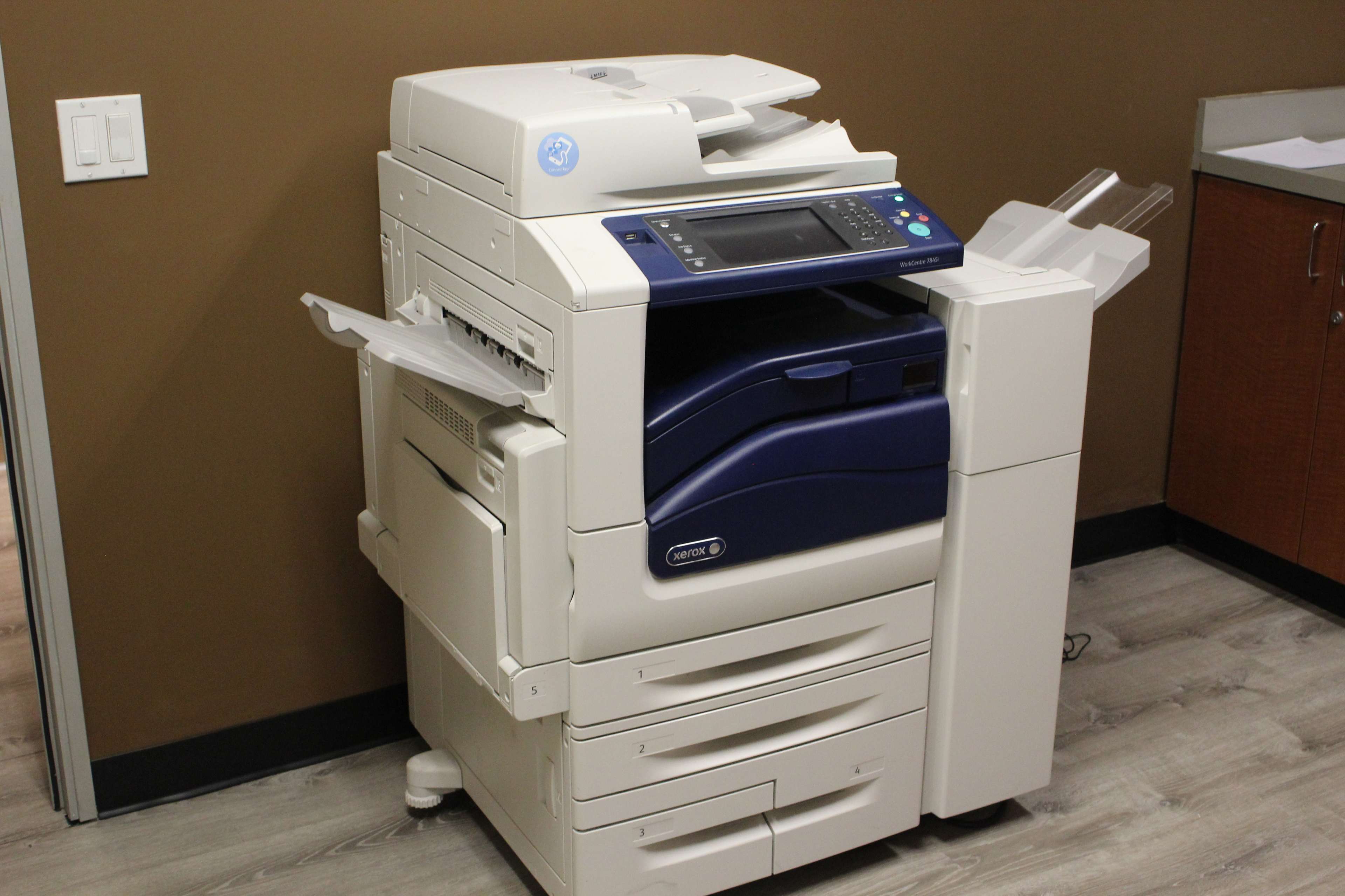 A multi-function printer is positioned in a well-lit office space with brown walls and wooden cabinets.