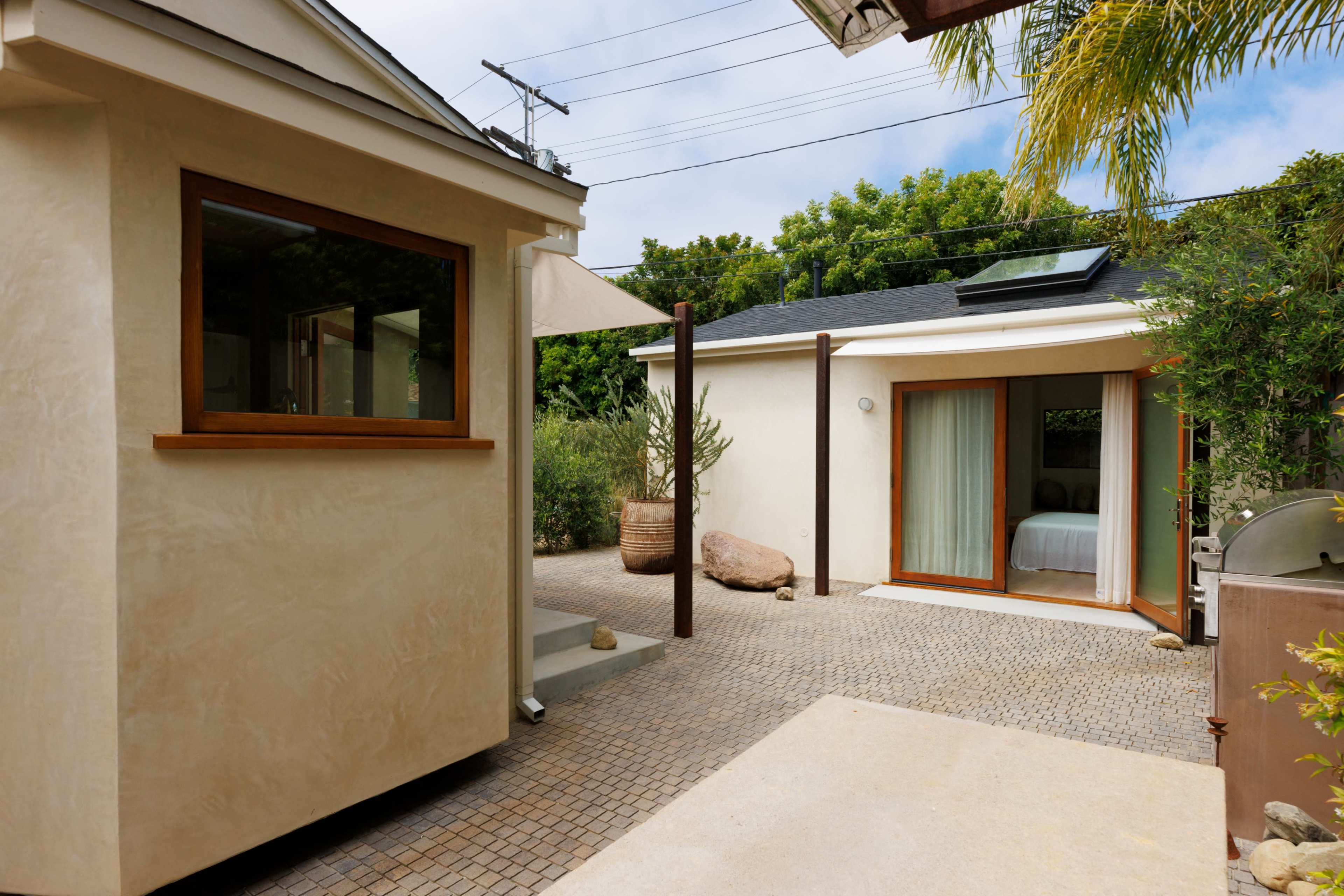 A small outdoor area featuring a smooth paved pathway with a light-colored cottage on the left and a larger room with large windows straight ahead, surrounded by greenery.