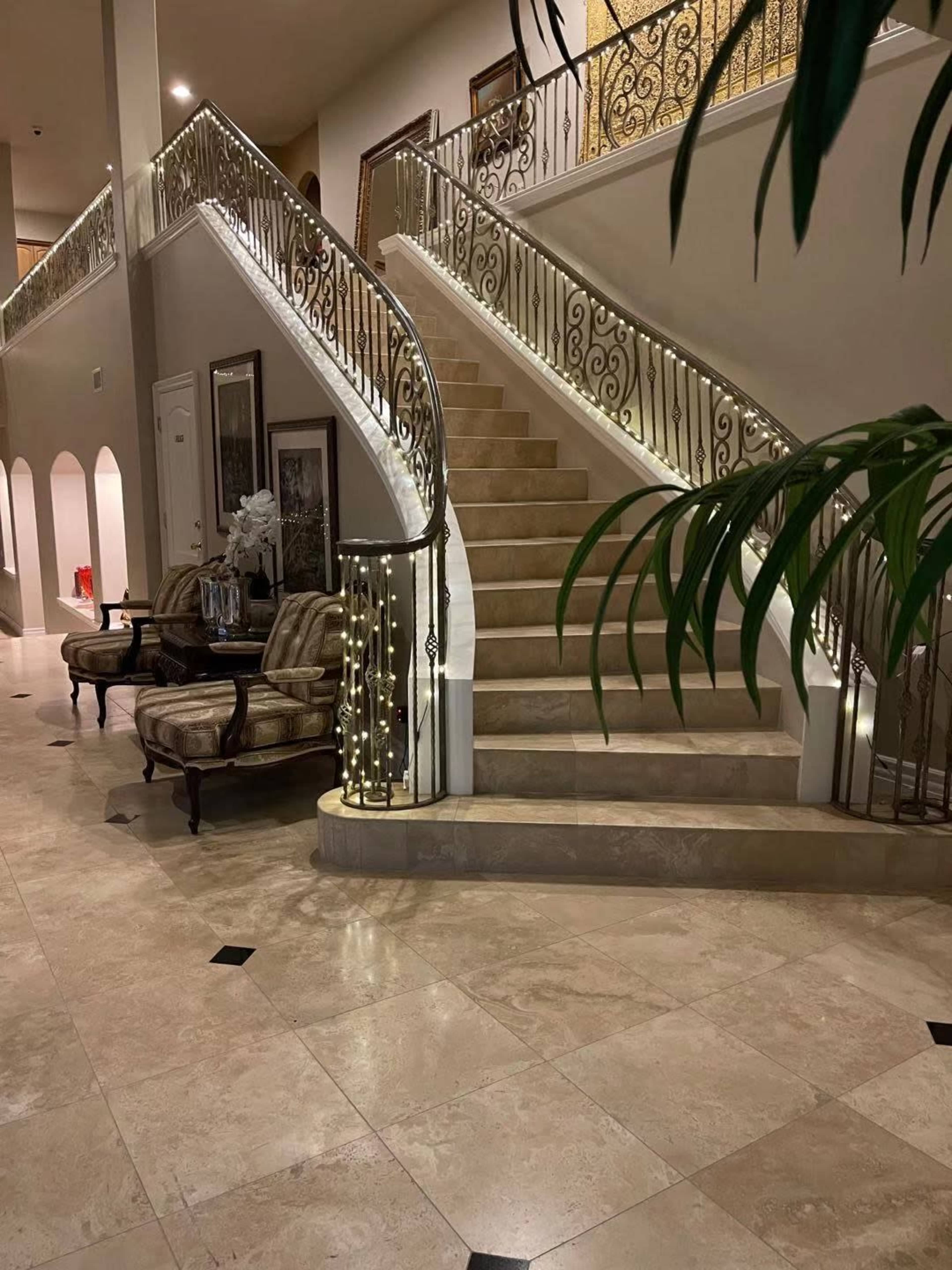A spacious entryway featuring an elegant staircase with a decorative railing, surrounded by marble floors and a pair of upholstered chairs.
